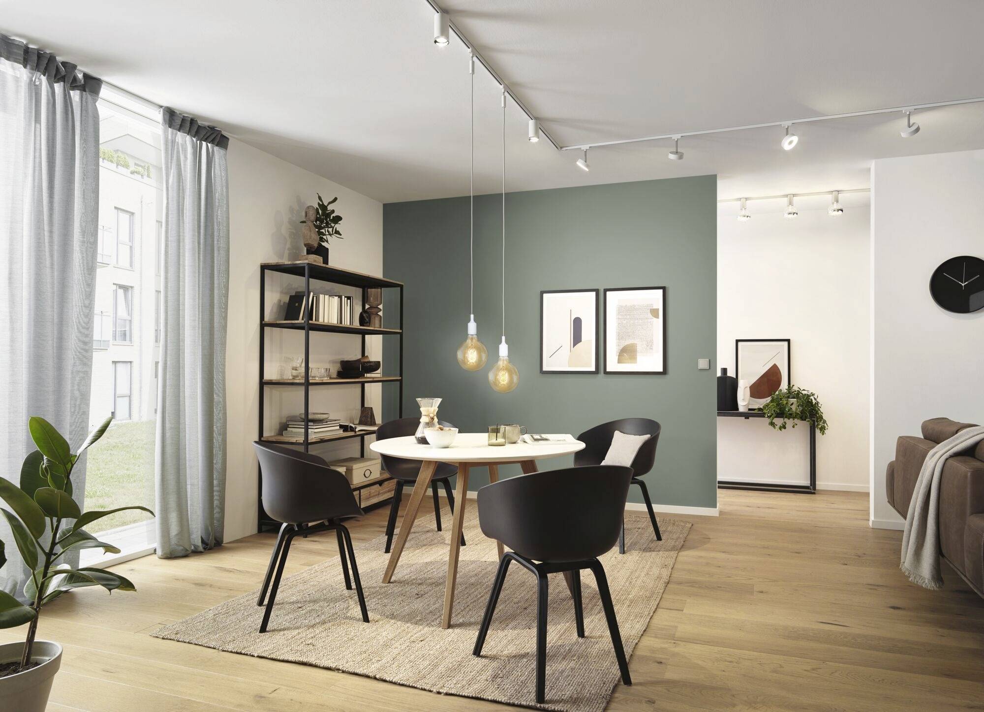Modern dining area with a round wooden table, four black chairs, a light green accent wall, and hanging lamps. Window allows daylight to enter.