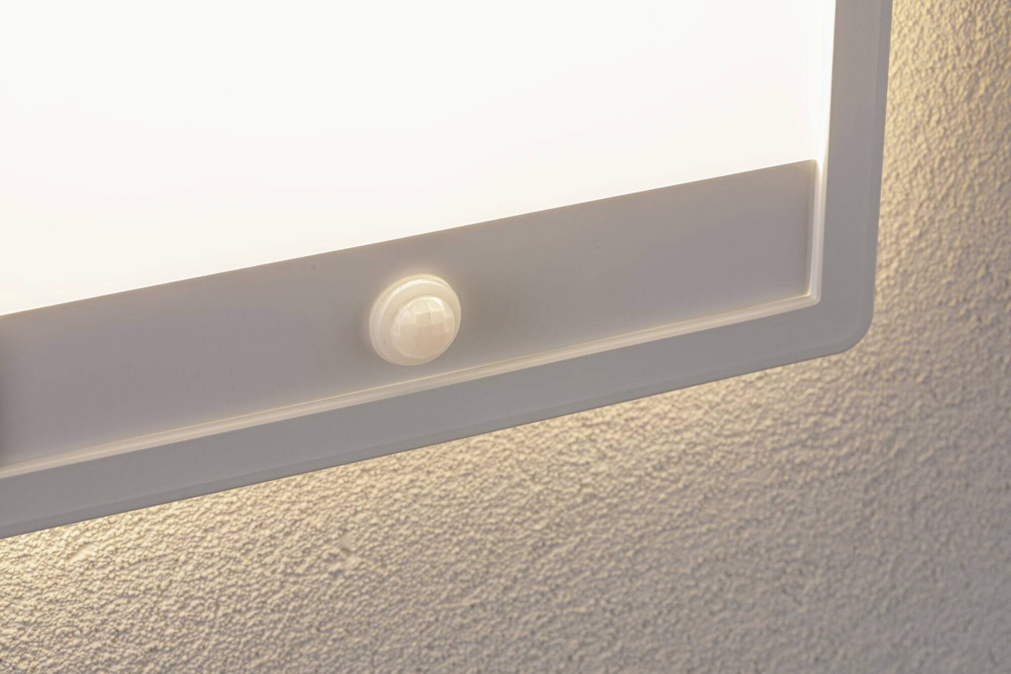 A white lamp with a motion sensor is mounted on the wall. It shines brightly and illuminates the surroundings evenly.