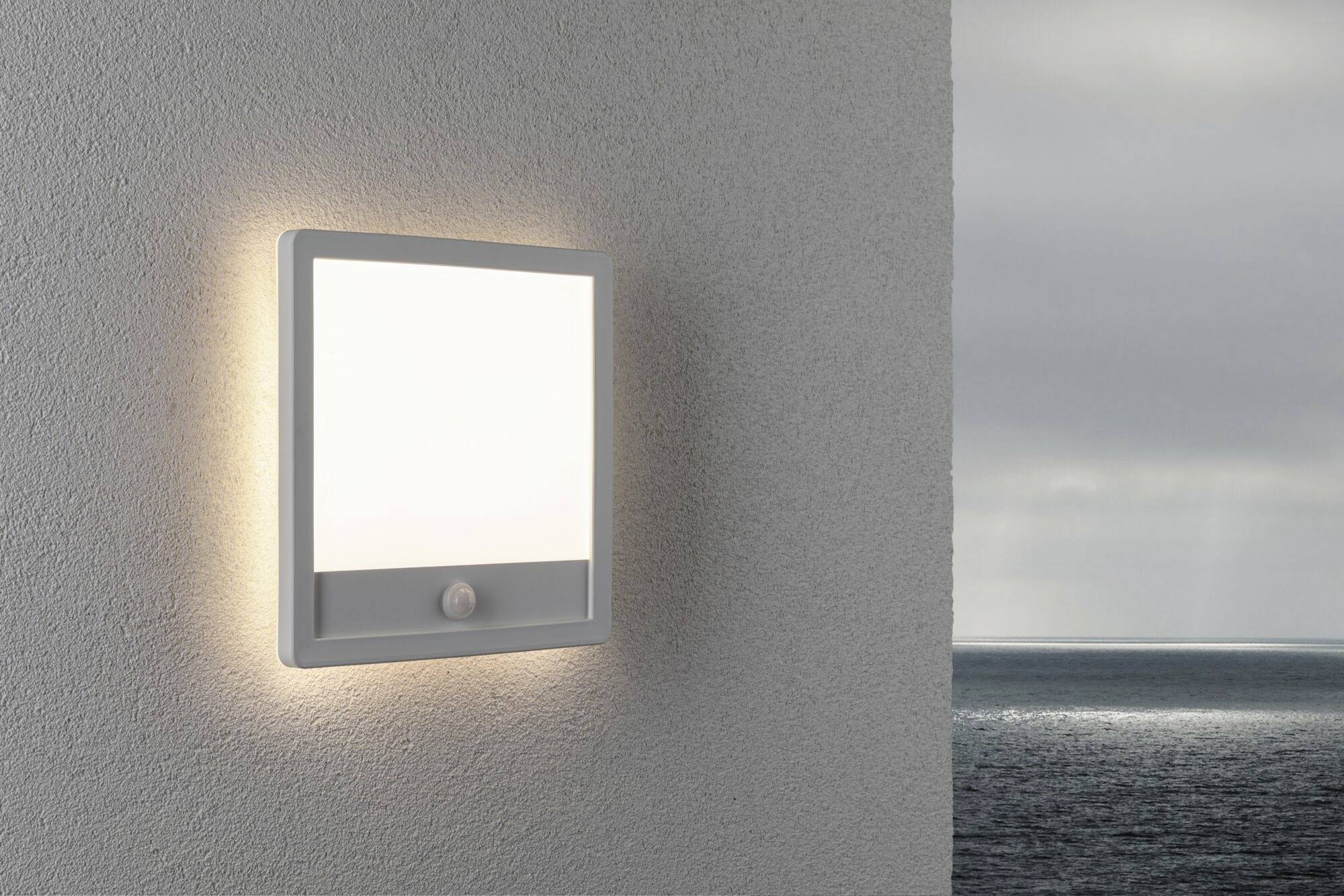 An exterior lamp illuminates a wall with a view of the sea. The sky is cloudy. The lamp features a simple, modern design.