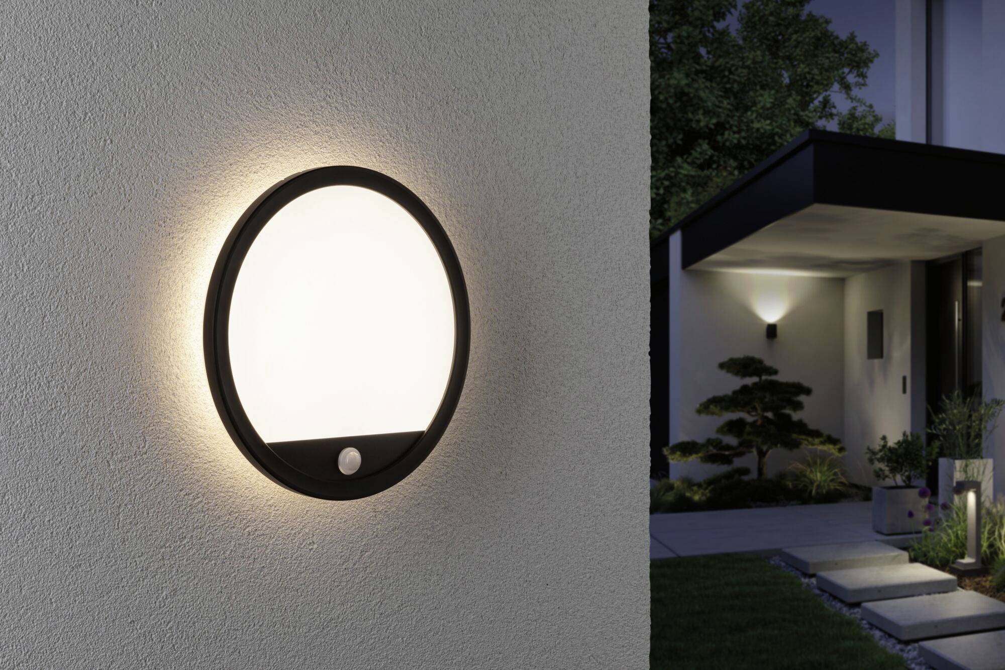 Round exterior wall light mounted on a house wall, emitting soft light. In the background, an illuminated house entrance and garden are visible.