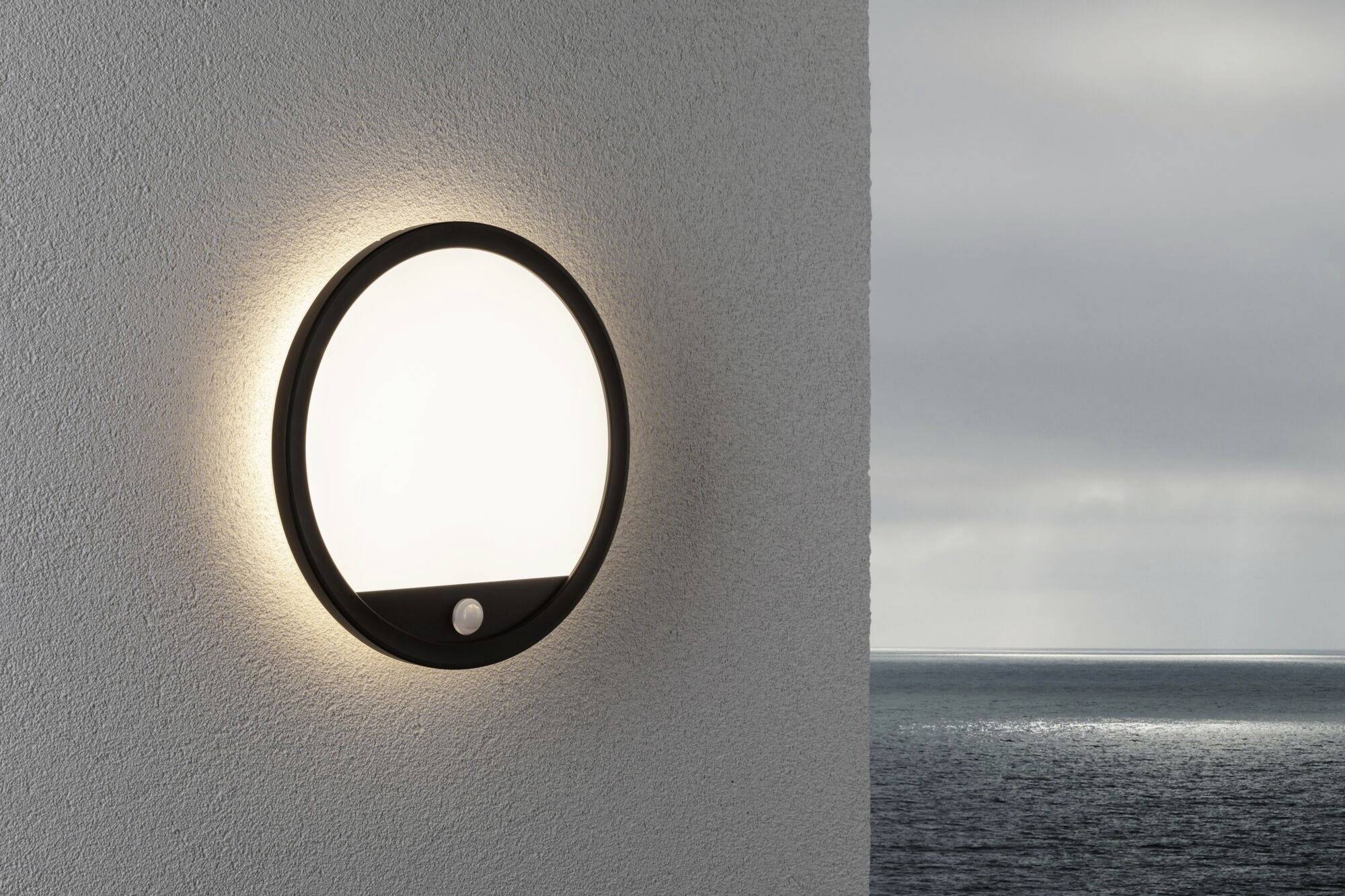 Wall light mounted on a white wall with a circular design and motion sensor; emits warm light. In the background, a cloudy sky over the sea.