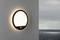 Wall light mounted on a white wall with a circular design and motion sensor; emits warm light. In the background, a cloudy sky over the sea.