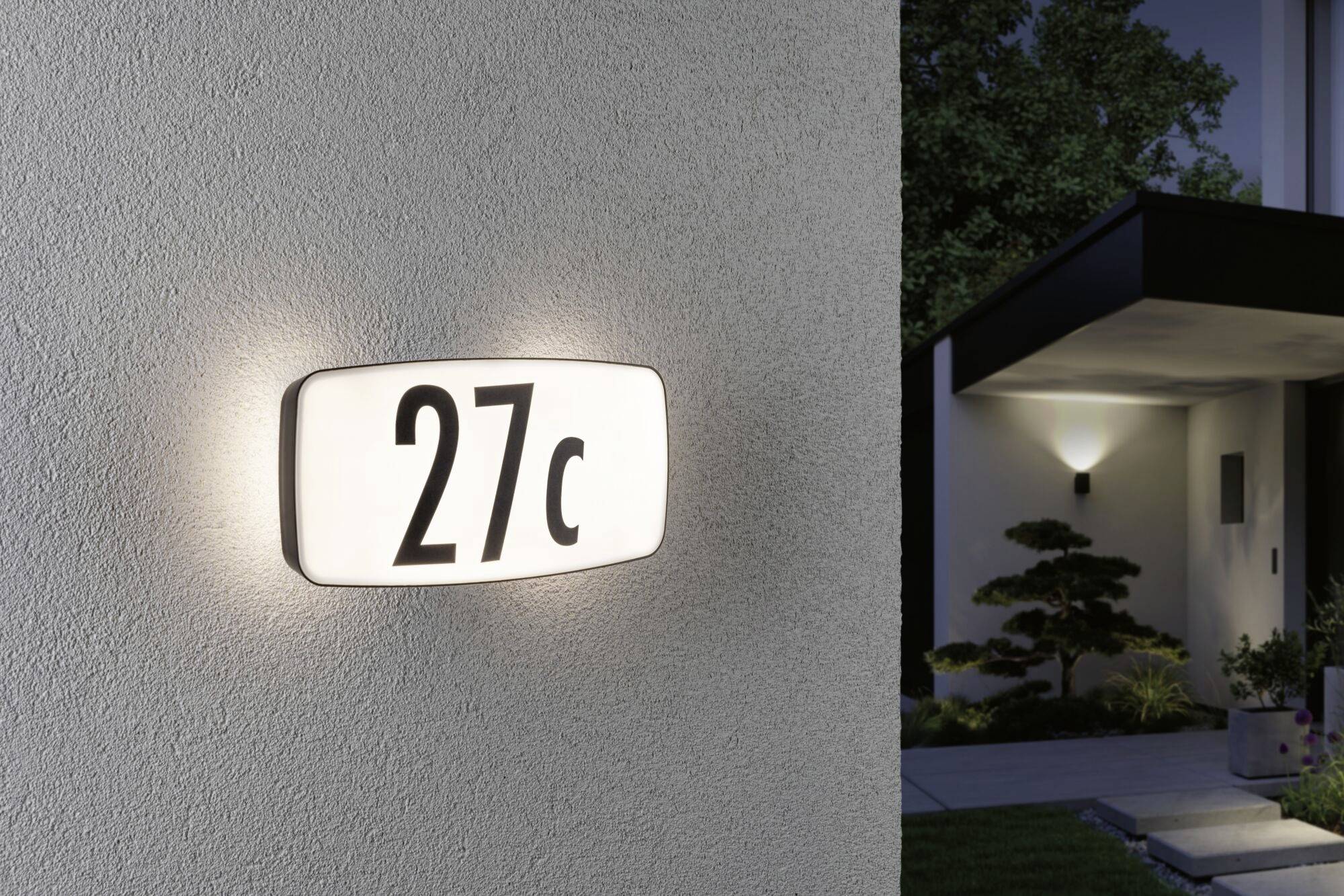 House number '27c' glows on the wall of a modern building, illuminated by a wall light. In the background, a lit entrance area is visible.