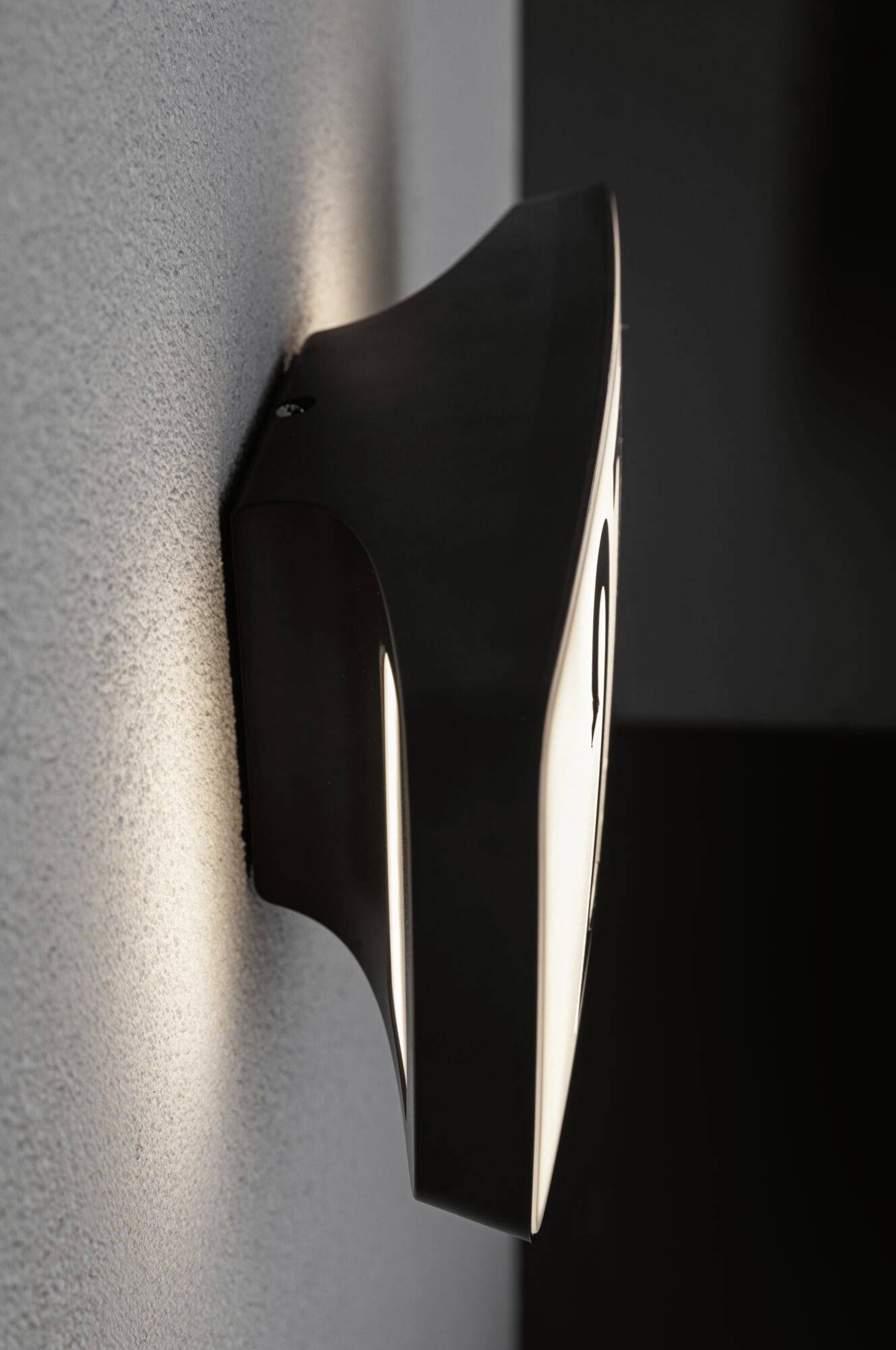 Black, contemporary wall light with indirect lighting, mounted on a grey wall, emitting a warm, diffused light.