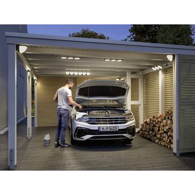 A man is checking the engine of a white car in an open carport. A stack of firewood is piled up beside it.