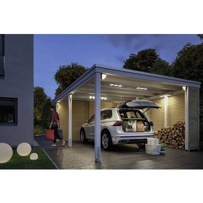 A person is carrying luggage to a lit, covered carport with an open boot filled with shopping.