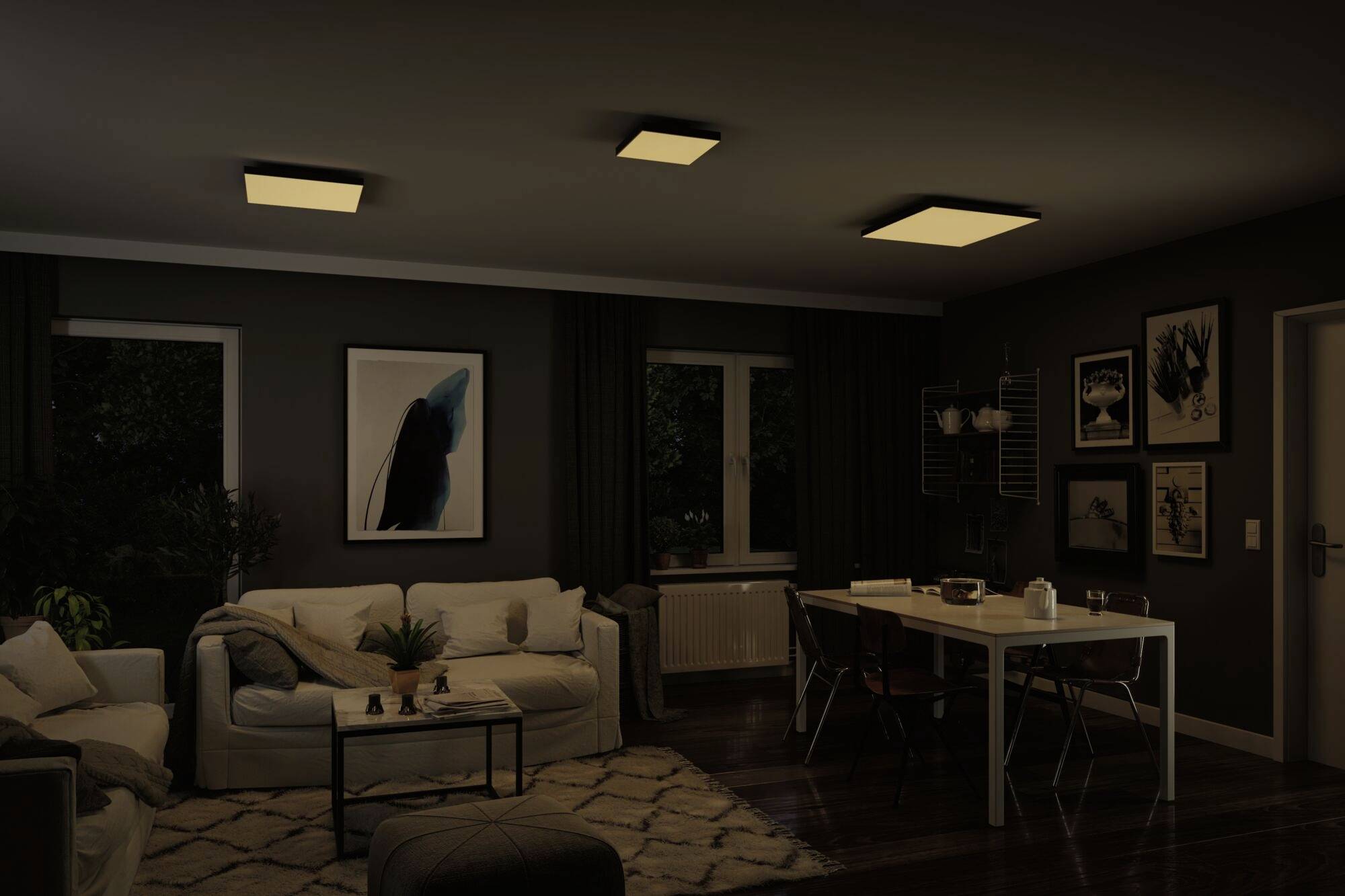 A living room with dim lighting. A sofa is situated on the left, a dining table with four chairs on the right. Artworks are hung on the walls.