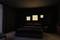 A bedroom with a large bed, dark bedding and bedside tables. Four illuminated square pictures are hanging on the wall.