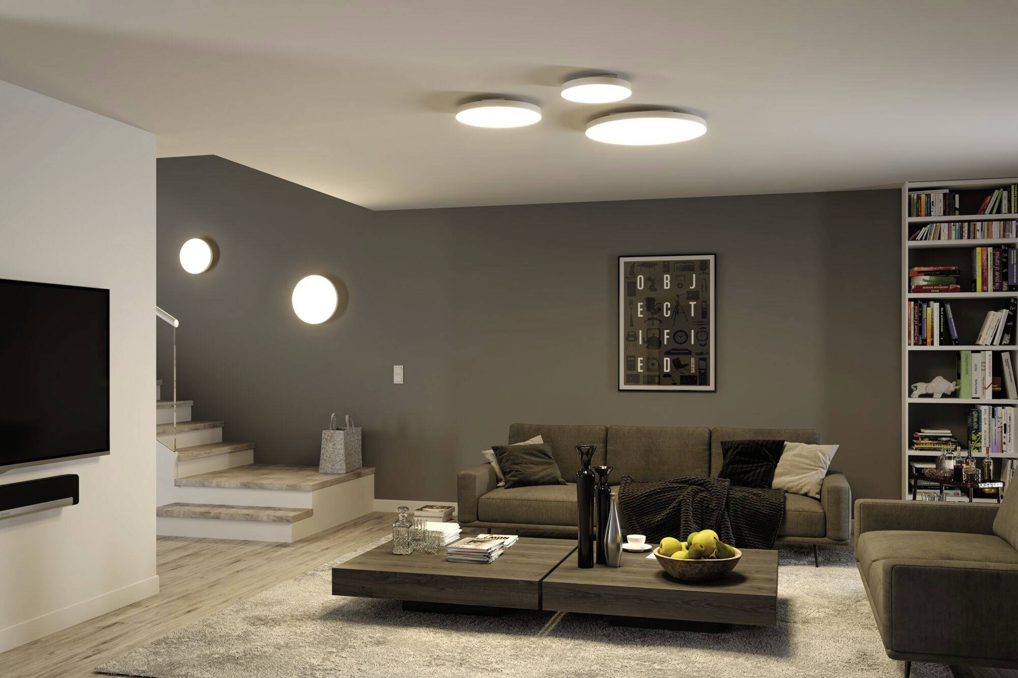 A modern living room with a grey sofa, coffee table, bookshelf and stairs in the background. Paintings and lights are hung on the wall.