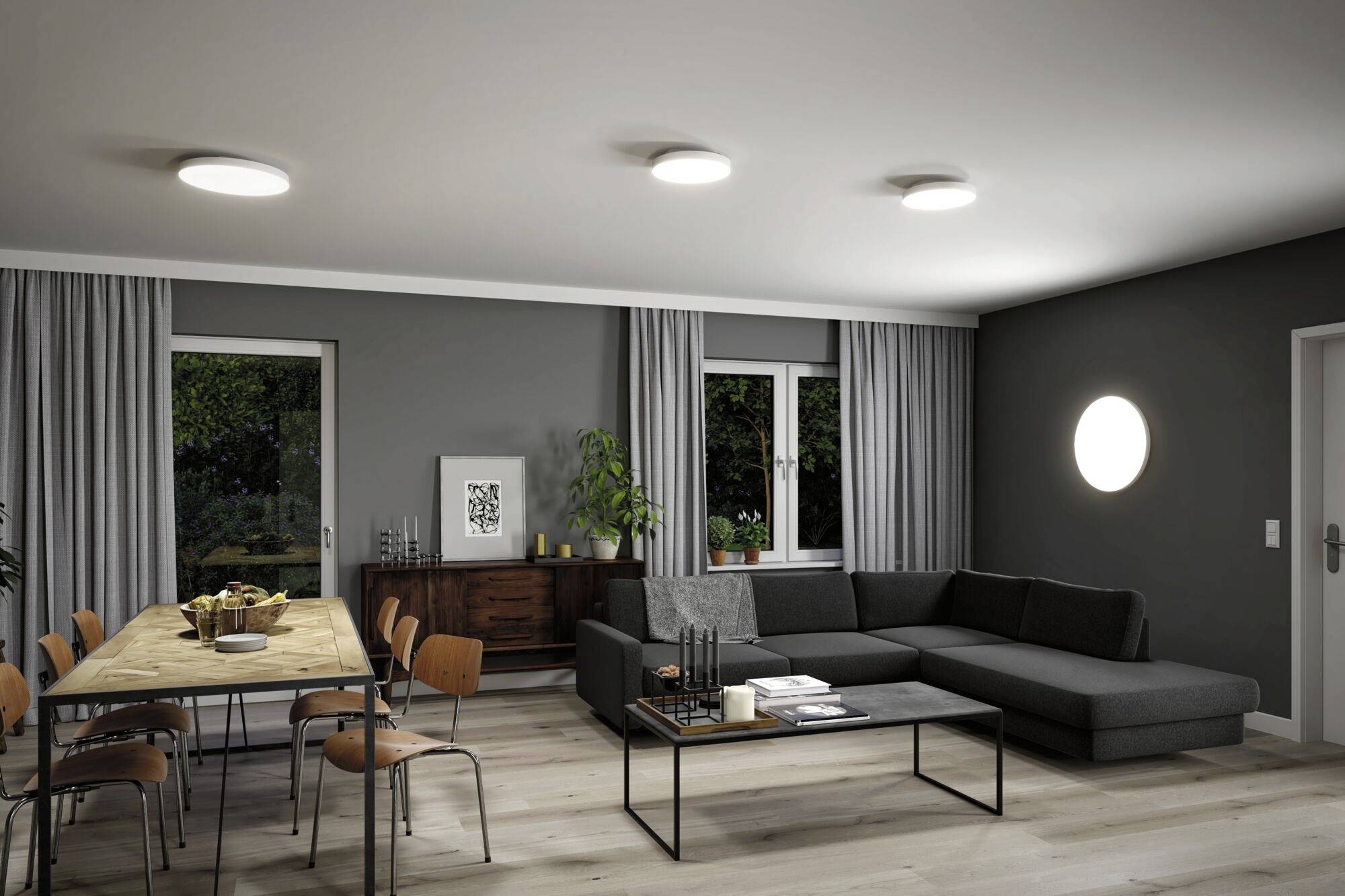 A modern living room with a grey sofa and a dining table for six people, featuring fine wooden and metal elements, well-lit with ceiling and wall lights.