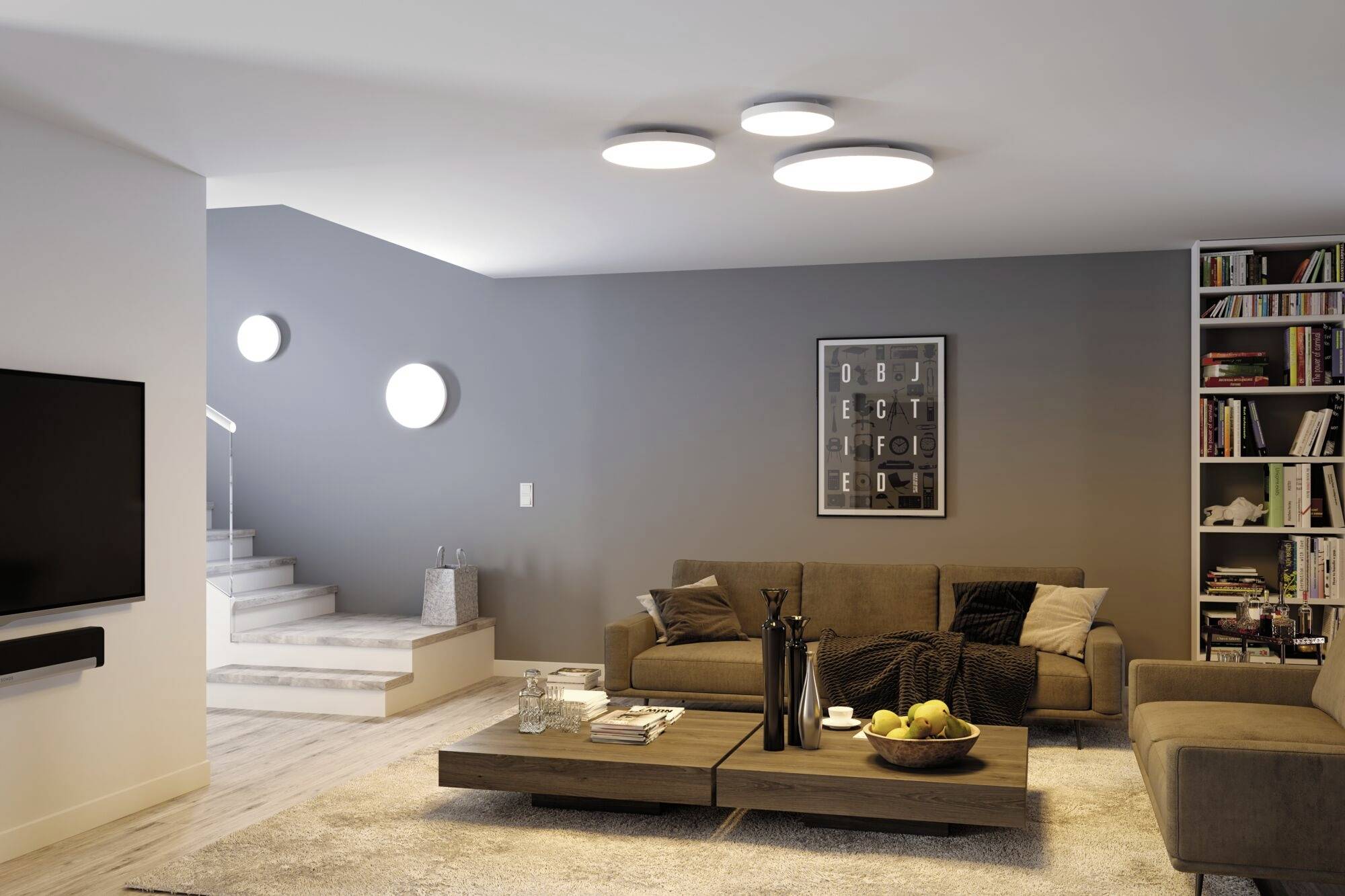 Living room with grey walls, modern sofa, coffee table with a fruit bowl, bookshelf full of books, wall art and staircase in the background.