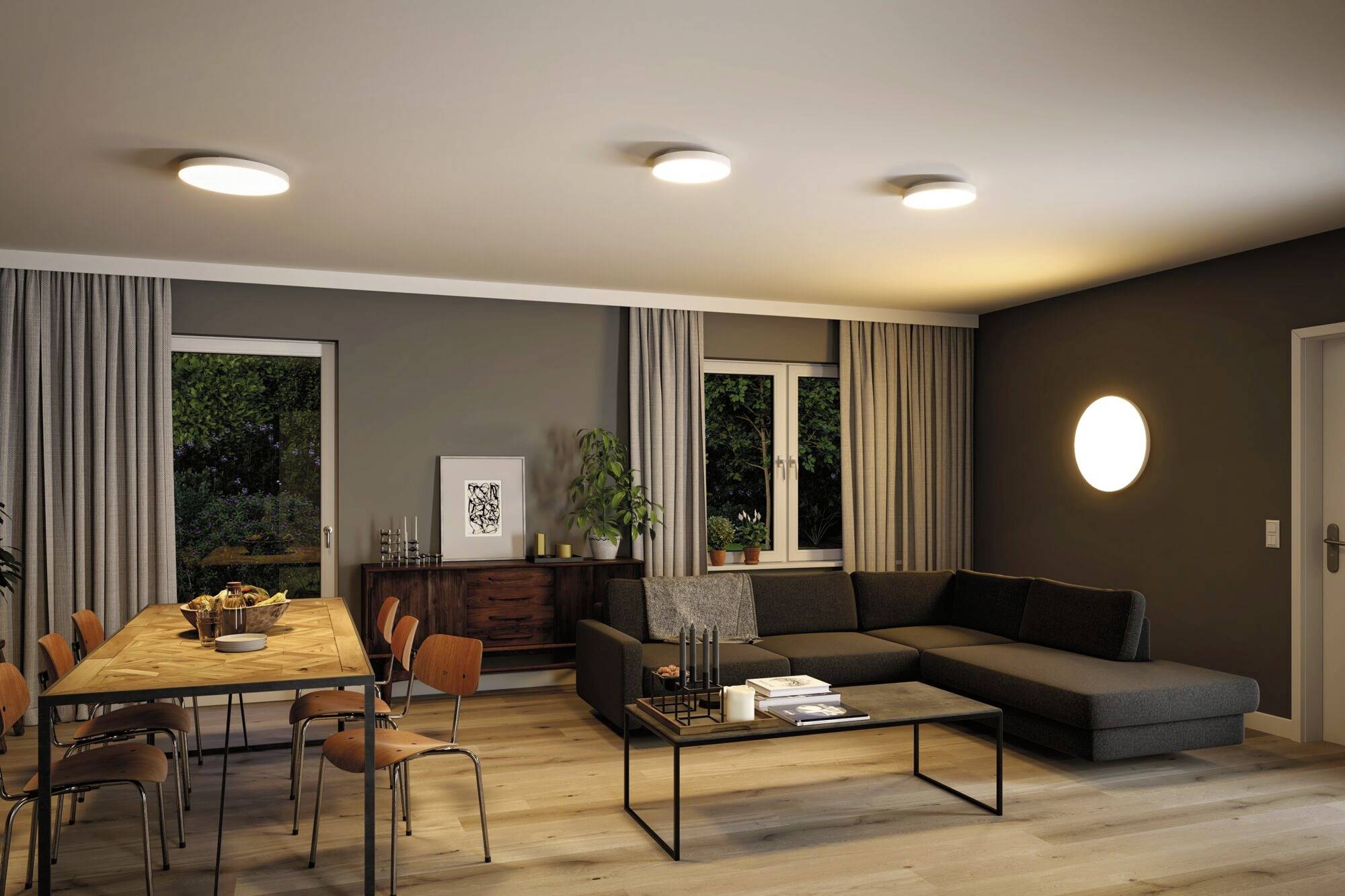 A modern furnished living room with a large corner sofa, a dining table with chairs, plants, and mood lighting.