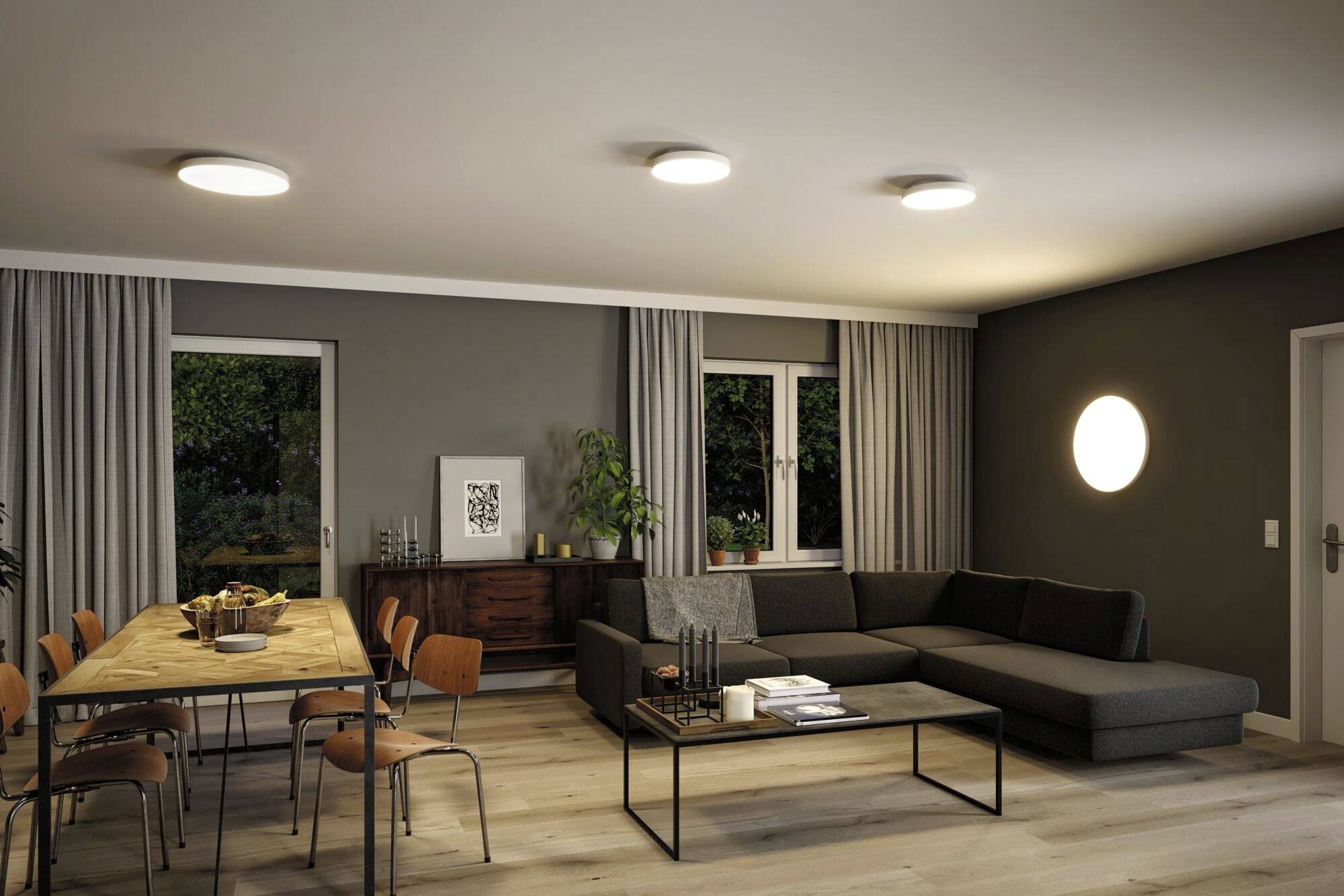 A modern living room with a large grey sofa, a coffee table and a dining table with chairs. On the right, a circular wall light.
