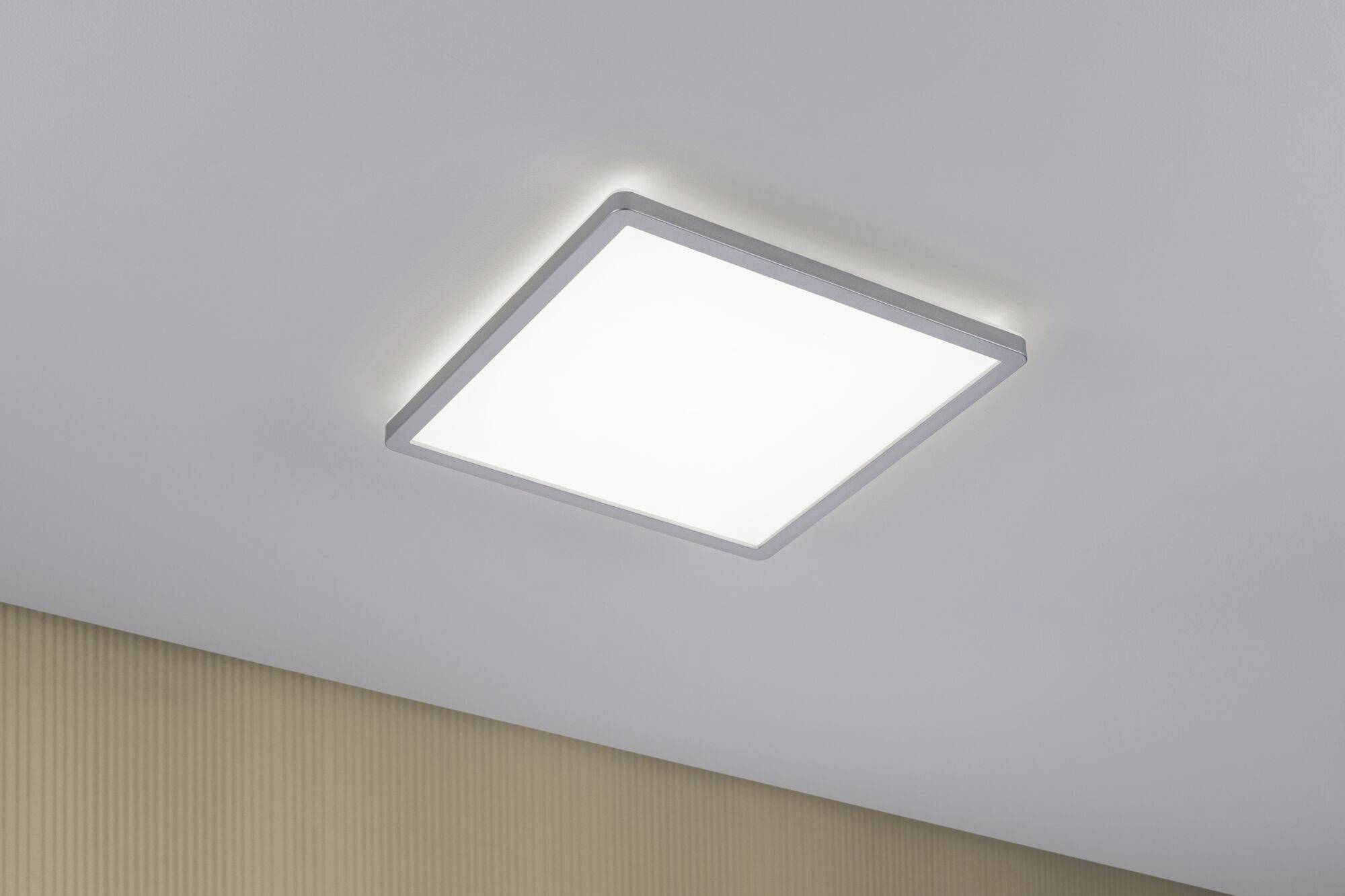 Square LED ceiling light, mounted on a white ceiling, emitting bright light. Beige wall in the background.