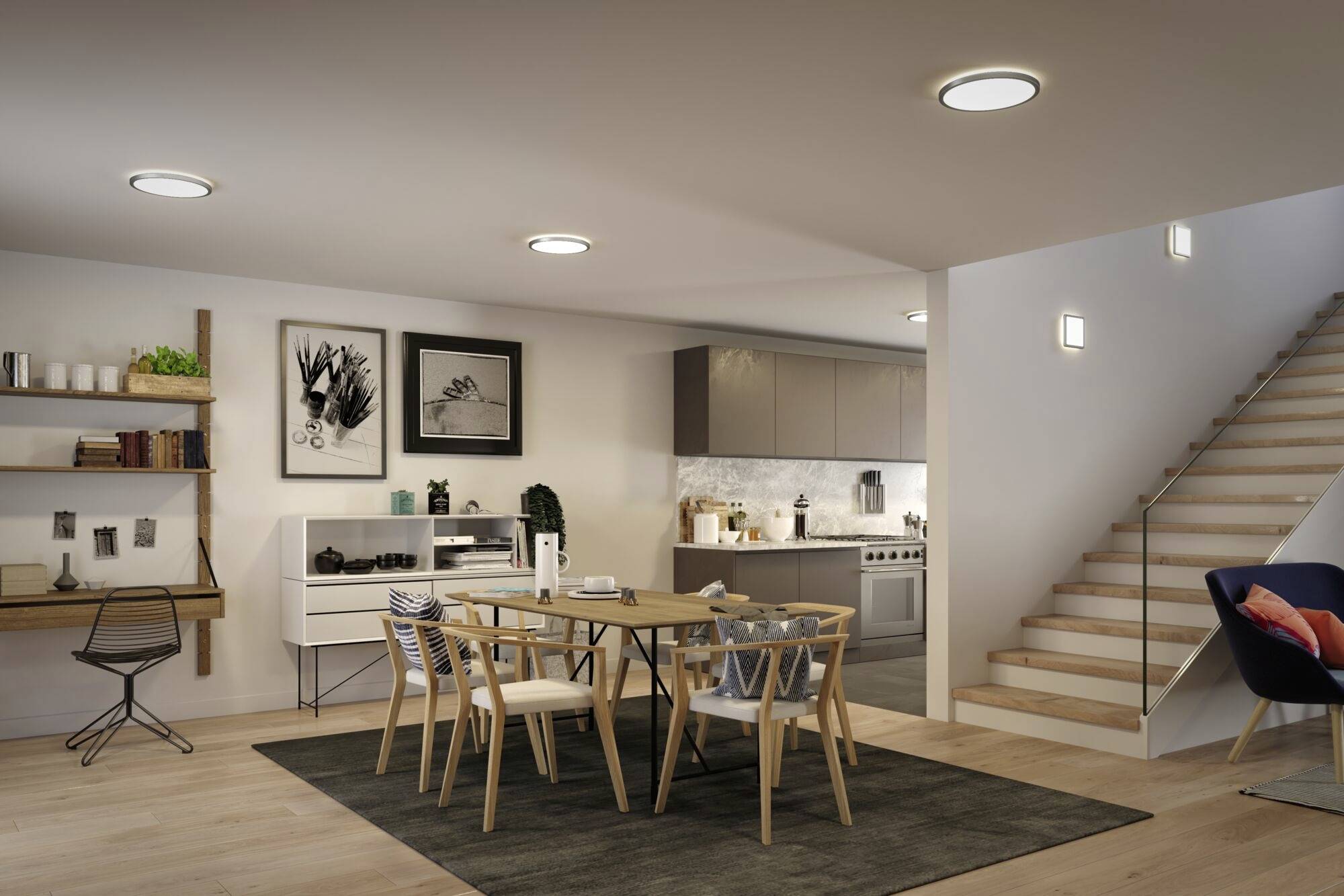 A contemporary dining room with a wooden table, six chairs and an adjoining kitchen. To the left, a wall-mounted shelving unit, to the right, a staircase, with warm lighting.