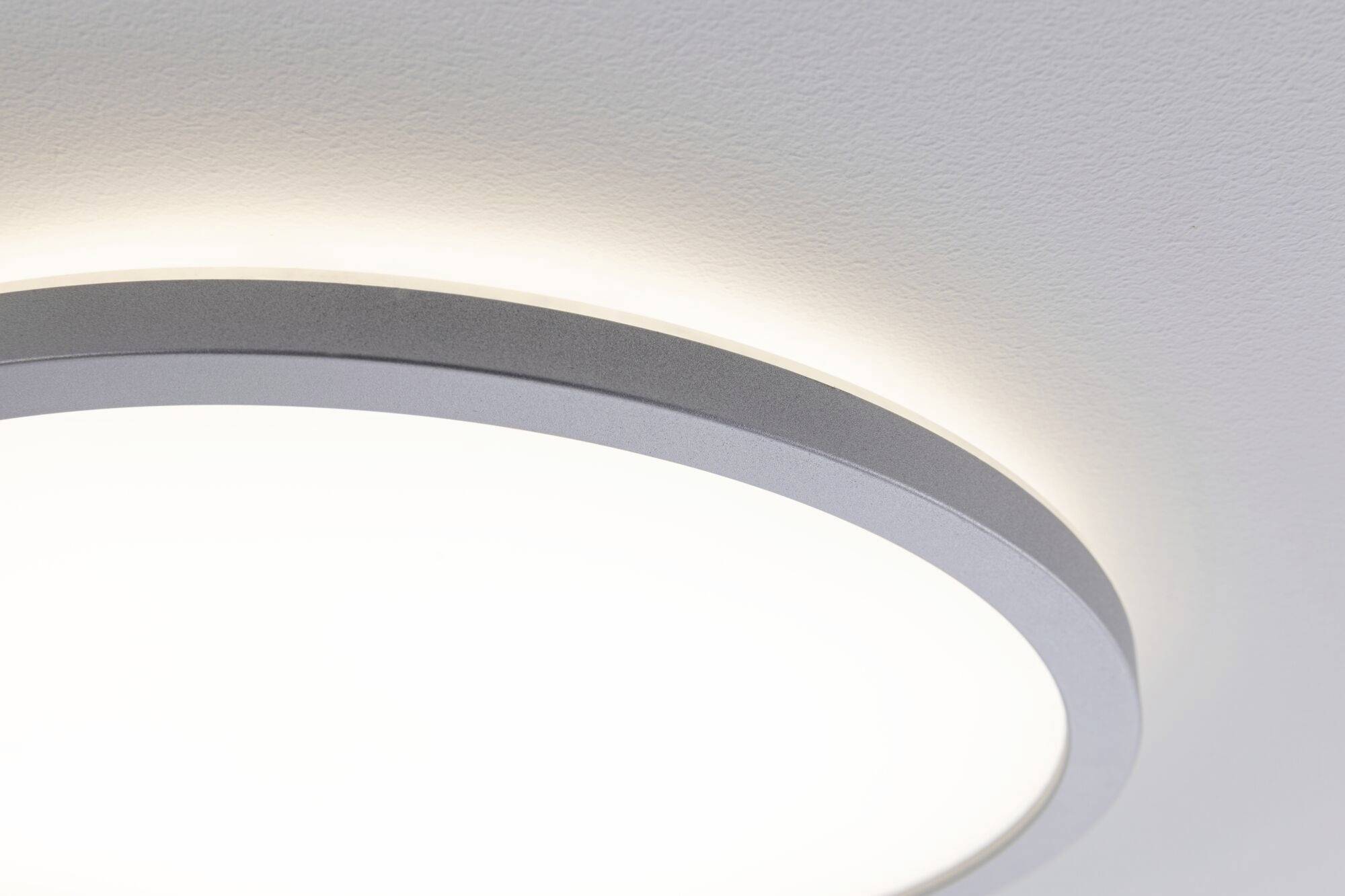 A round, modern ceiling light shines brightly against a white ceiling. The edges of the light are silver.