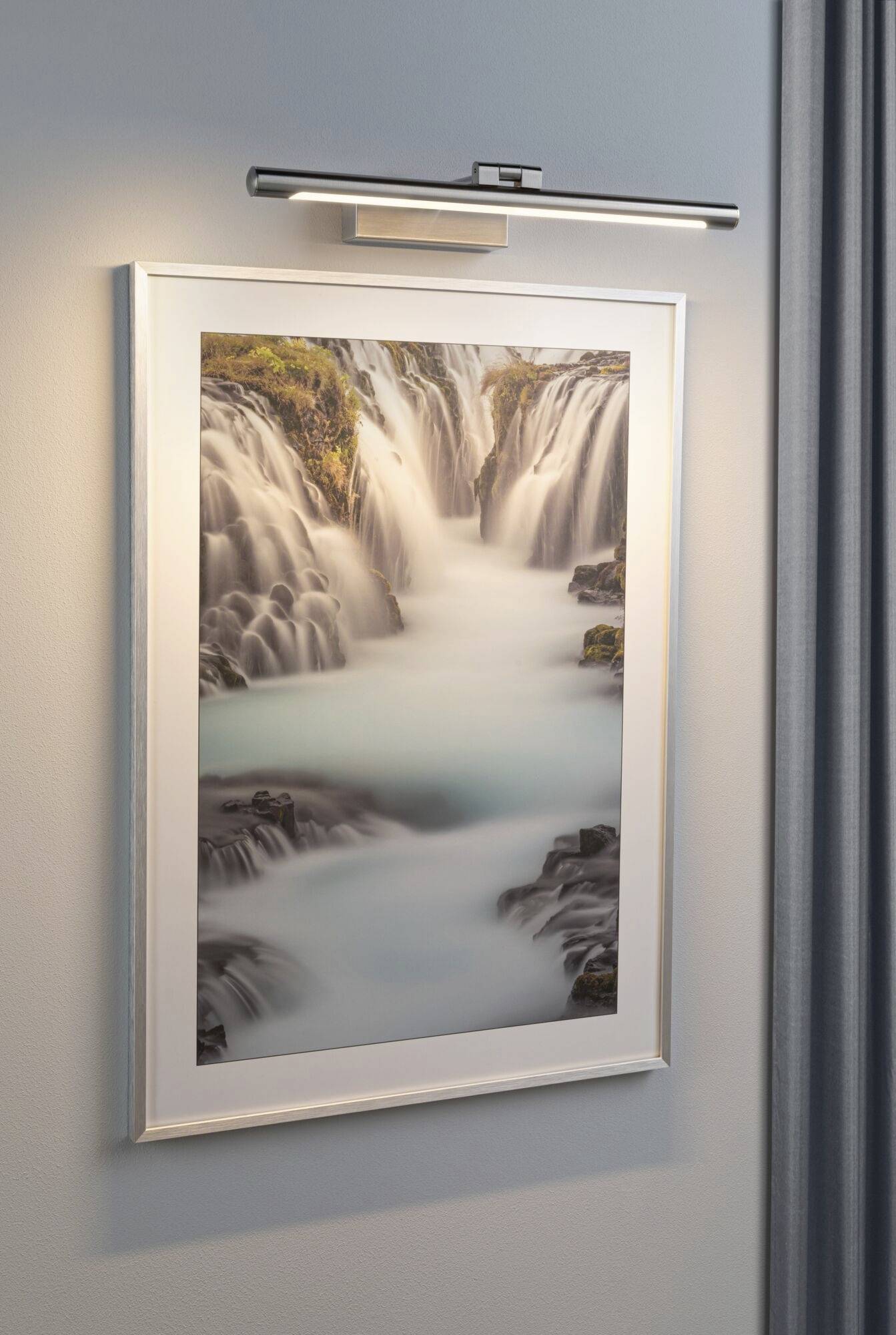 A framed picture of a waterfall hanging on a light grey wall, with a spotlight above it.