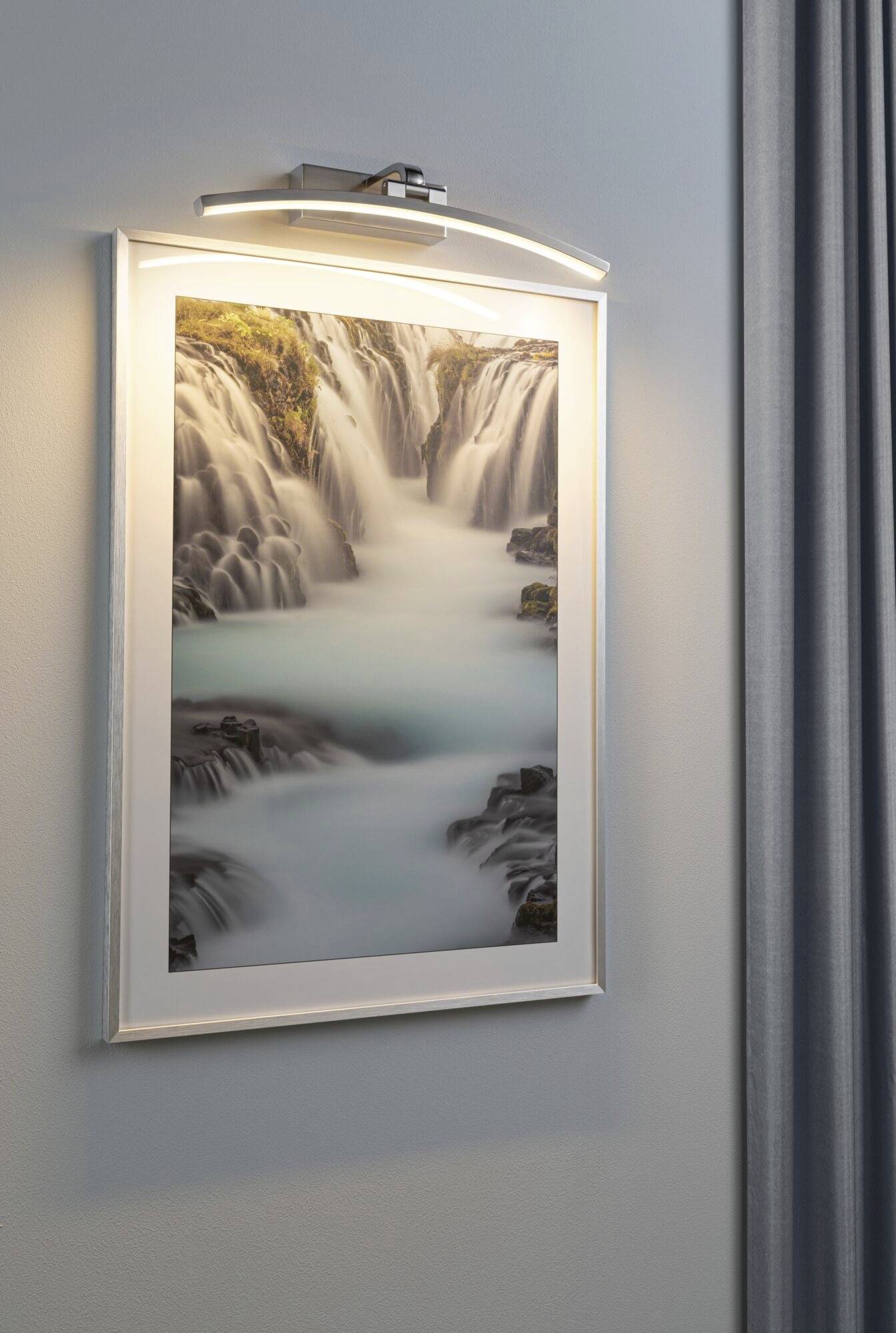 A picture of a waterfall hangs on a wall, illuminated by a lamp above it. The water appears soft due to a long exposure.