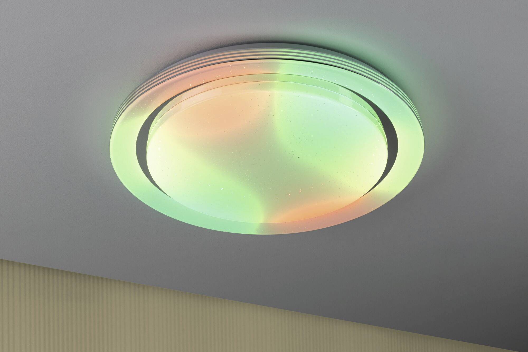 Round ceiling light with colour-changing LED lighting in green and orange. It is mounted on a grey ceiling.
