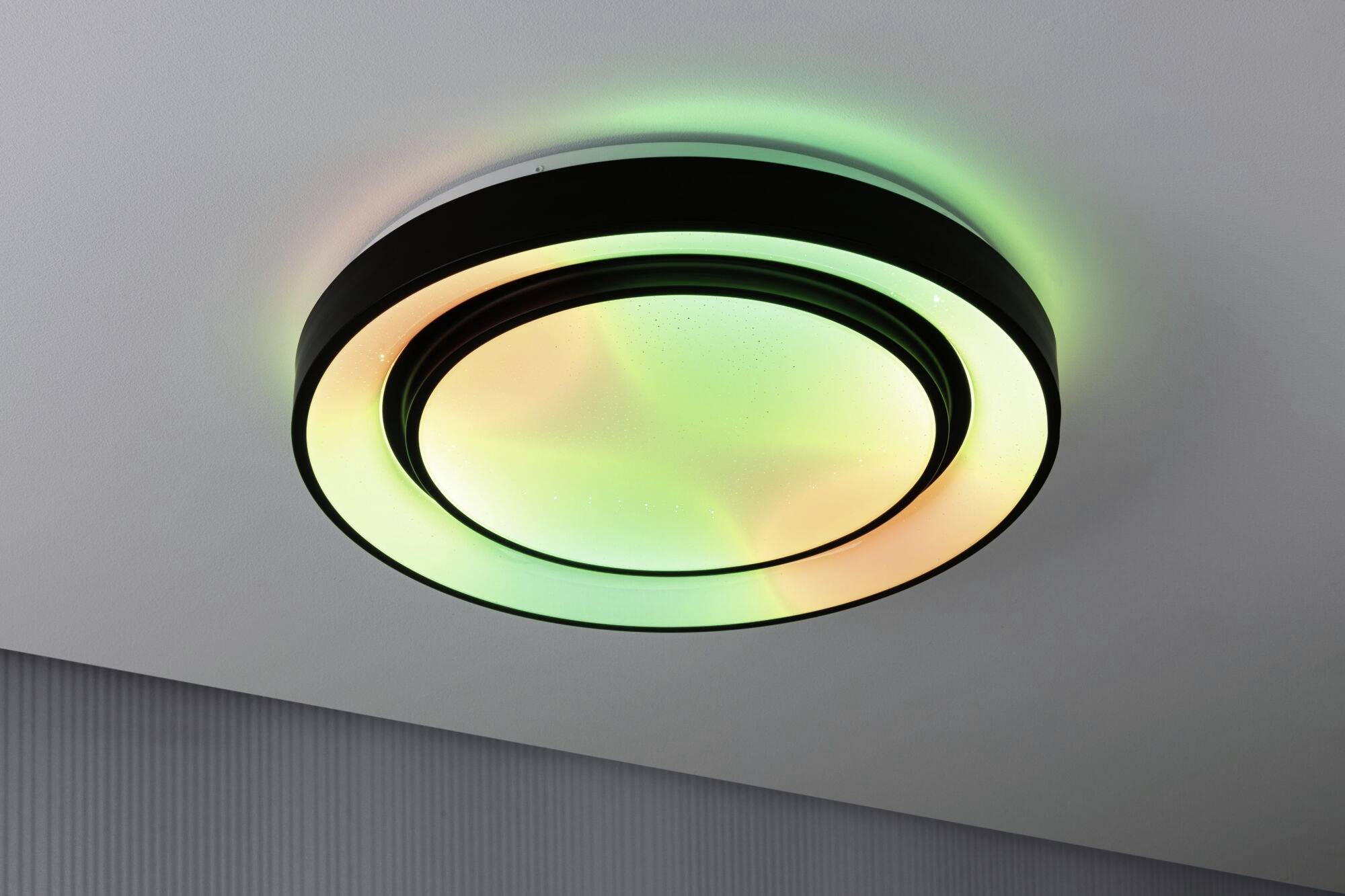 A round ceiling light with illuminated rings in green and orange hues, mounted on a white ceiling.