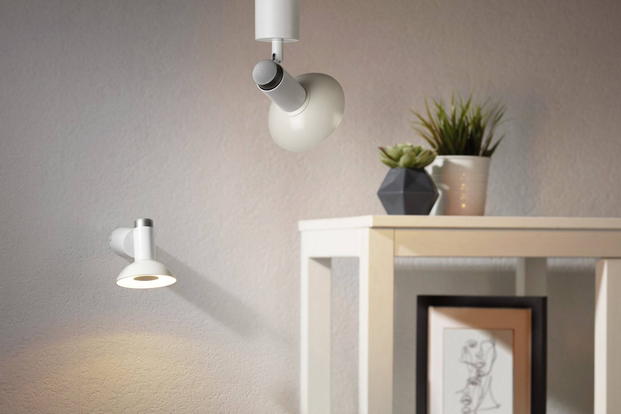 A white table lamp illuminates a table with plants and a framed picture. Another lamp is mounted on the wall.