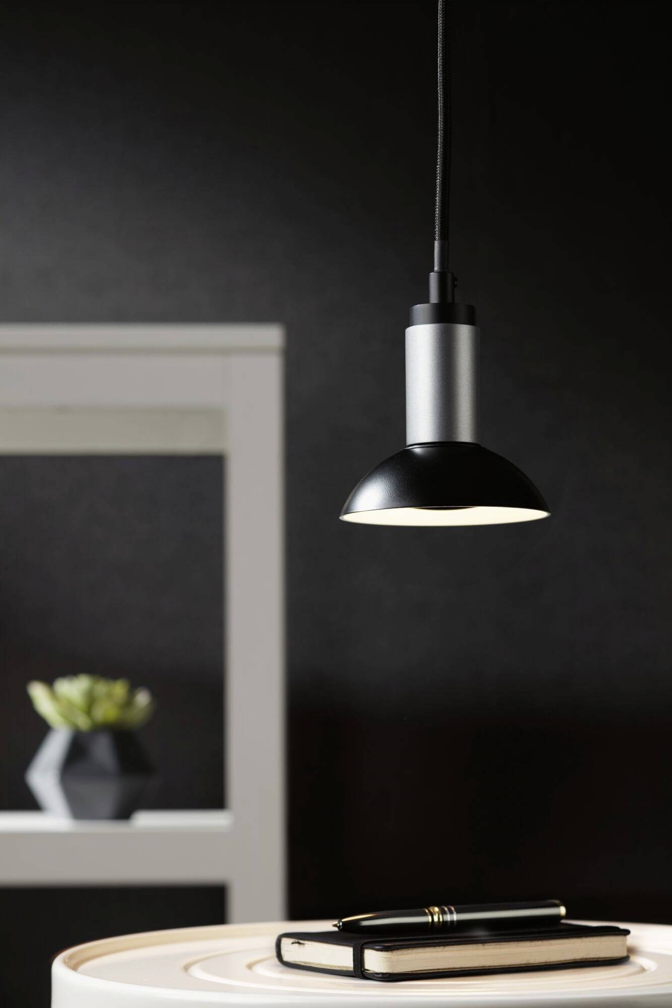A contemporary pendant light illuminates a table with a notebook and pen. A plant sits on a shelf in the background.