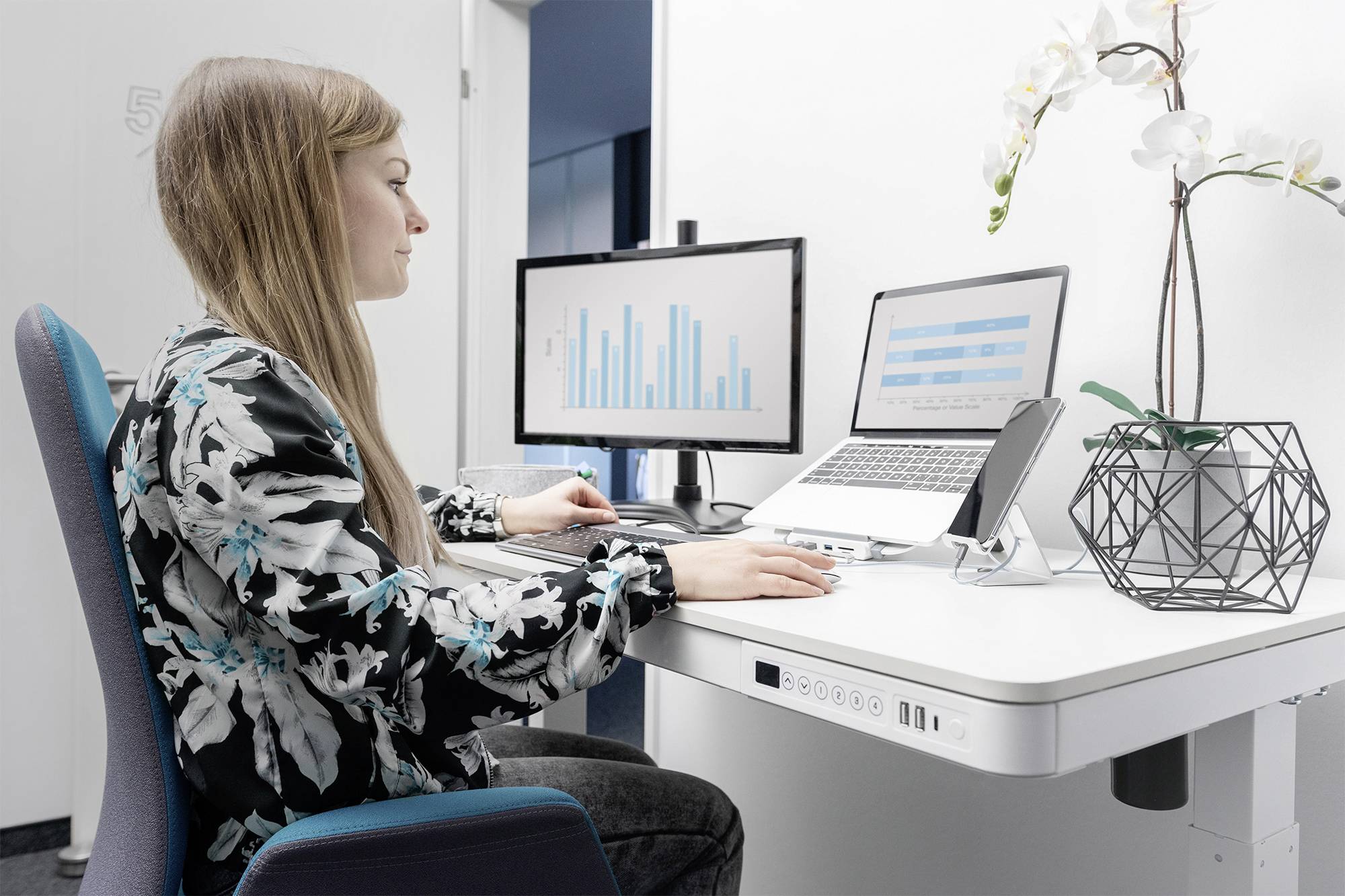 A woman is sitting at a desk working on a laptop. Bar graphs are visible on the monitor. An orchid is placed beside her.