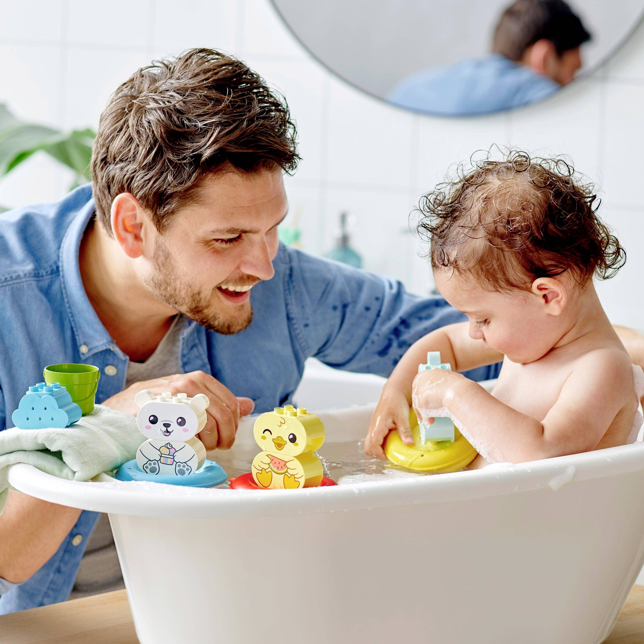 A man is playing with a toddler in the bath. They have colourful bath toys. Both are smiling and look happy.