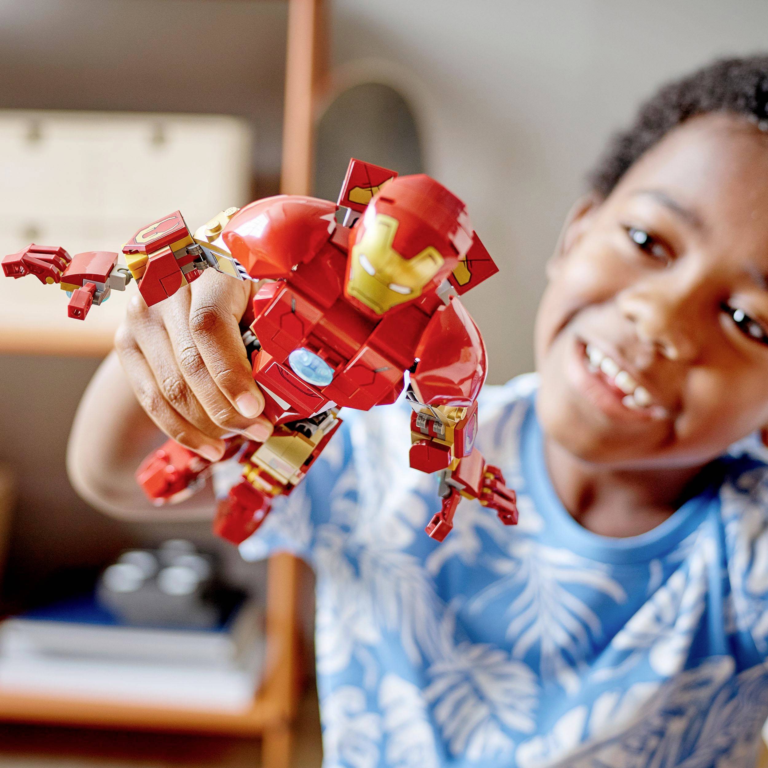 A child holds an Iron Man action figure enthusiastically towards the camera, with a bookshelf visible in the background.