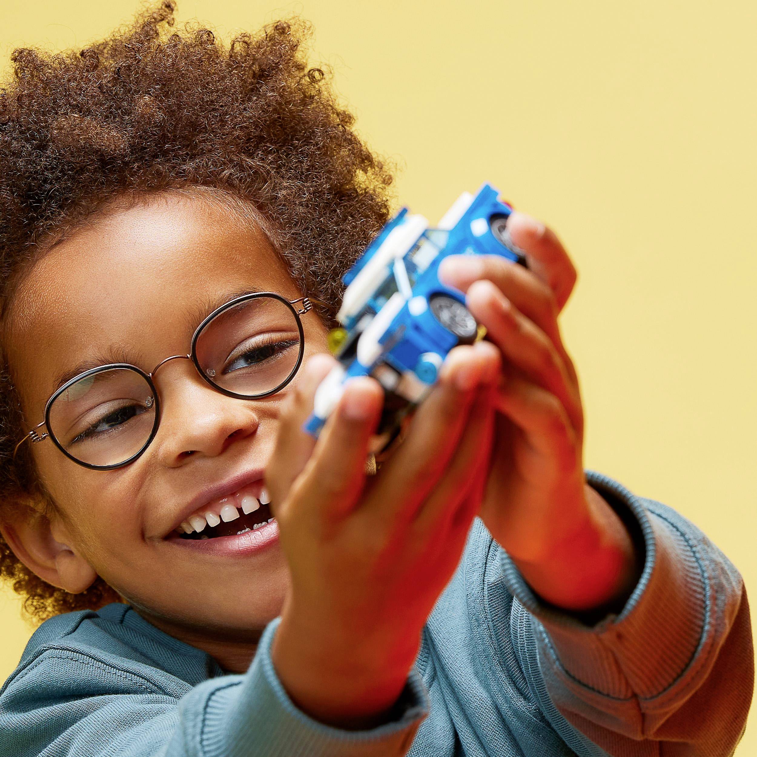 A child with curly hair and glasses smiles while playing with a toy car. Yellow background.