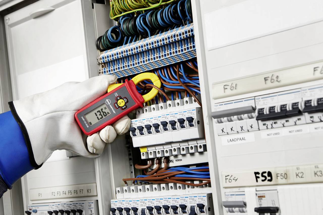 A person is measuring the electrical voltage in a control cabinet using a measuring device. The value displayed is 137.6.