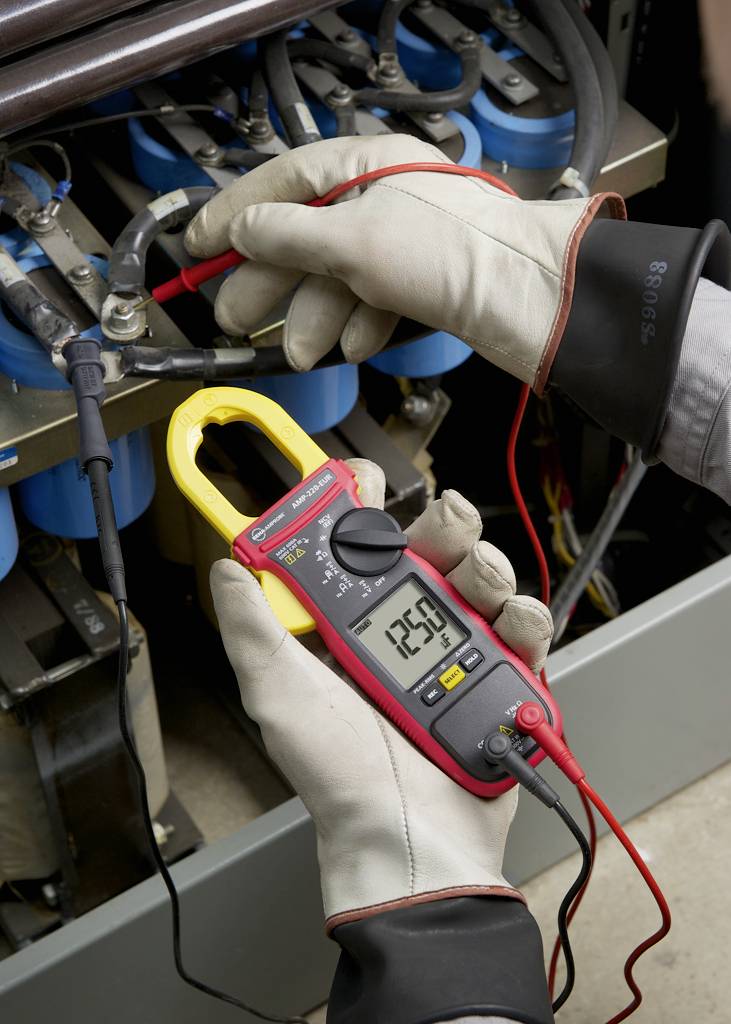 A person wearing gloves is measuring the electrical voltage with a digital multimeter across a row of blue batteries.