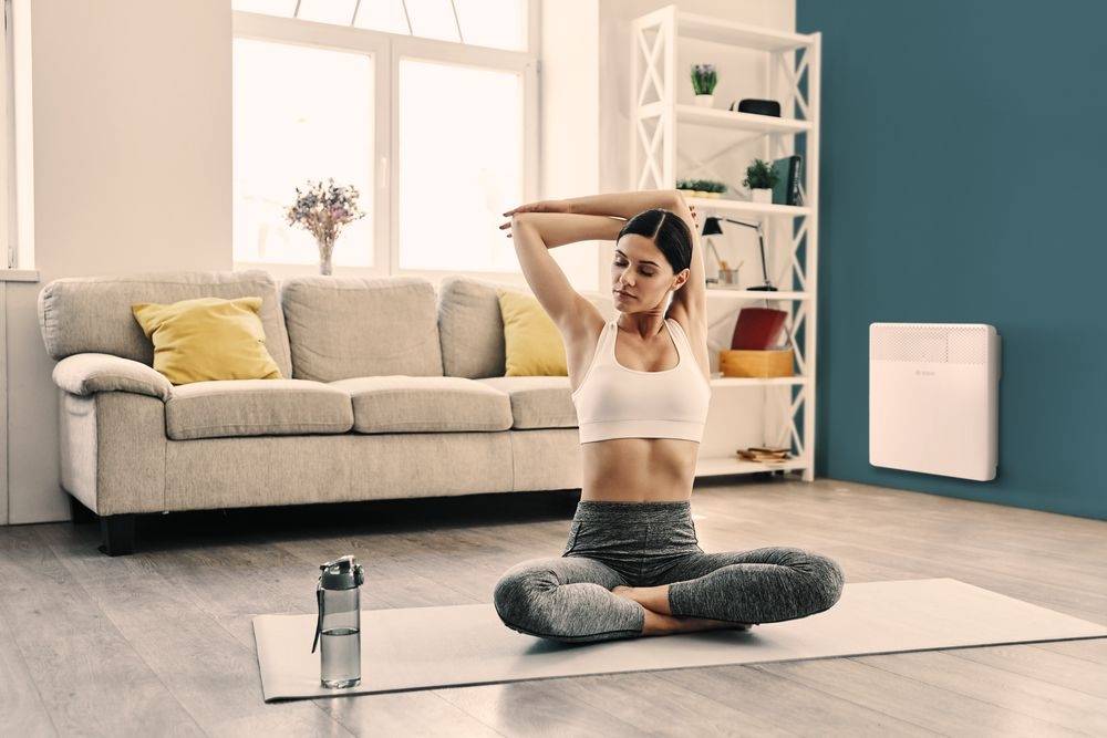 A woman is sitting on a yoga mat in a living room, stretching her arms. In the background, there is a sofa and a bookshelf.