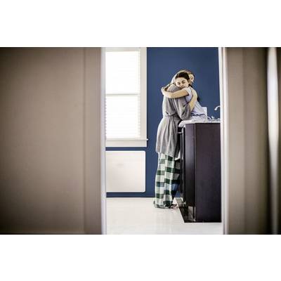 A person in pyjamas is hugging another person in the kitchen. They are standing near the sink. The background features a blue wall and a window.
