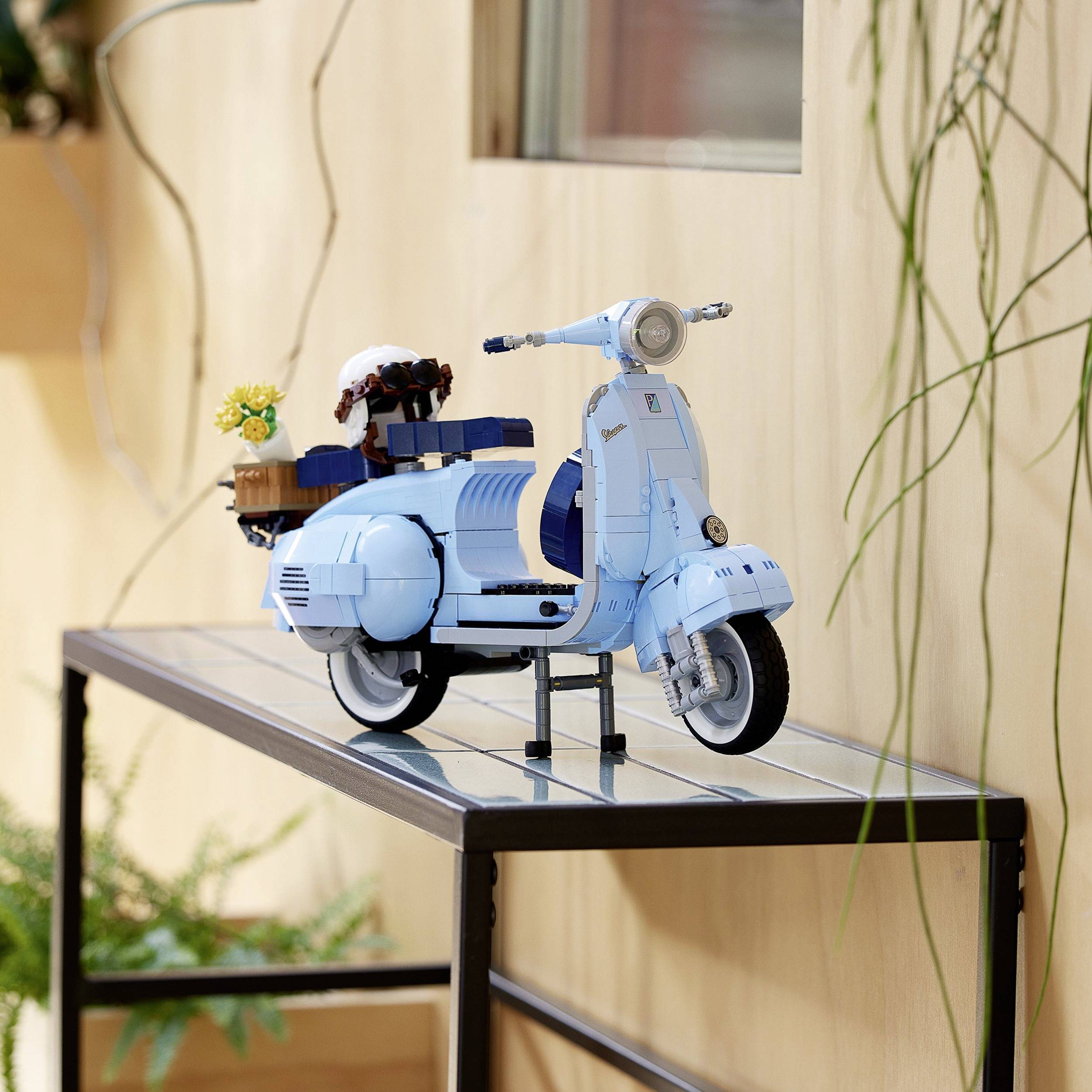 A model of a blue motor scooter is standing on a table. Beside the seat is a basket with flowers. Plants can be seen in the background.