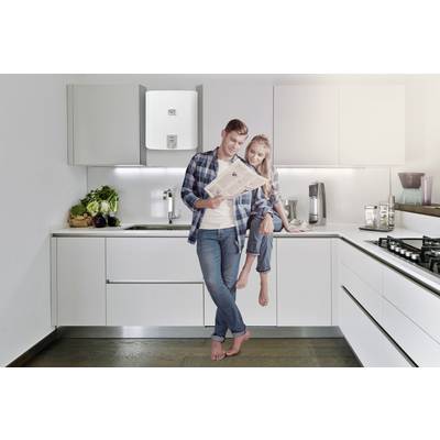 A smiling couple stands in a modern kitchen. The man is reading a newspaper, while the woman sits on the worktop. Fresh vegetables can be seen in the background.