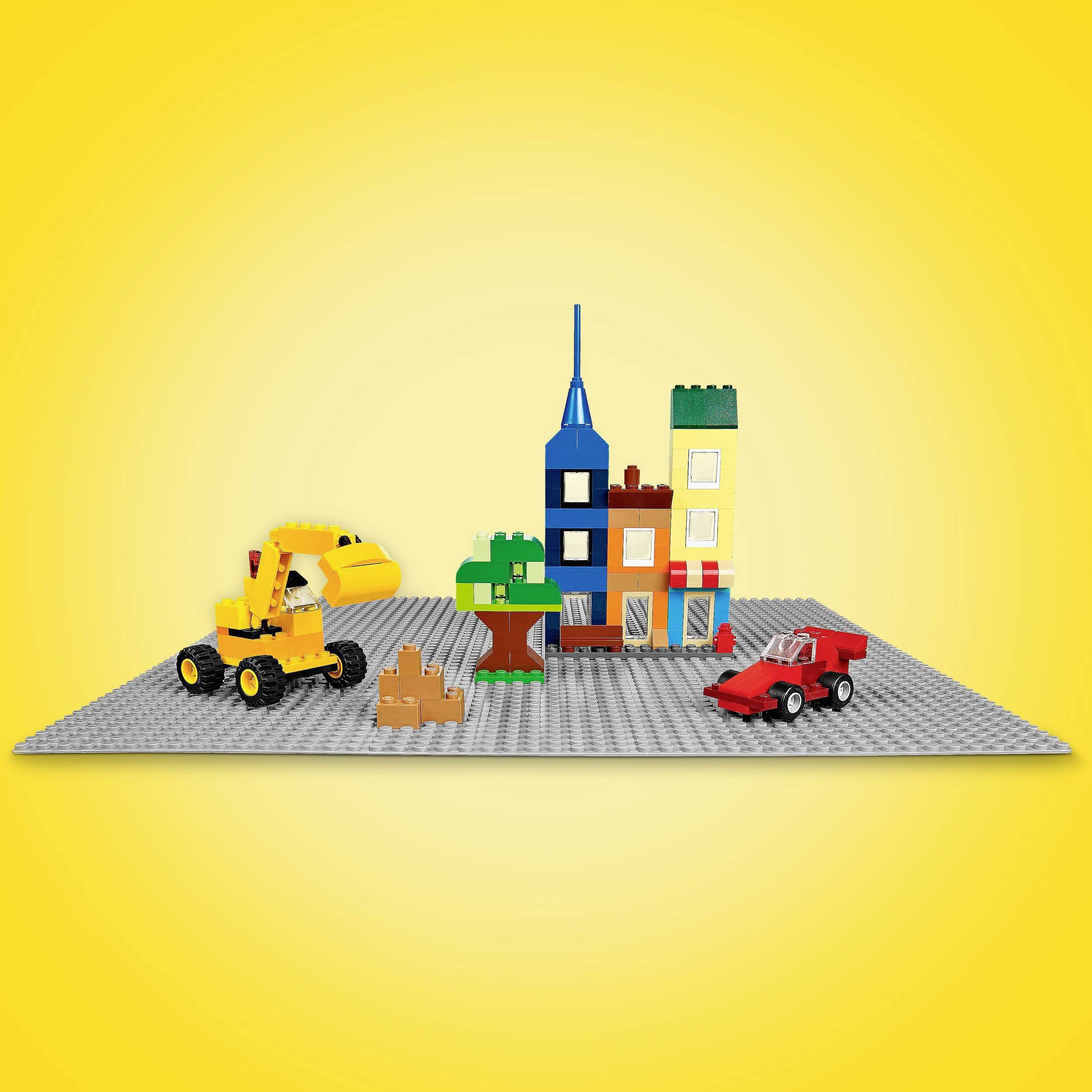 A digger and a red car made of building blocks in front of a colourful house and a tree on a grey base against a yellow background.