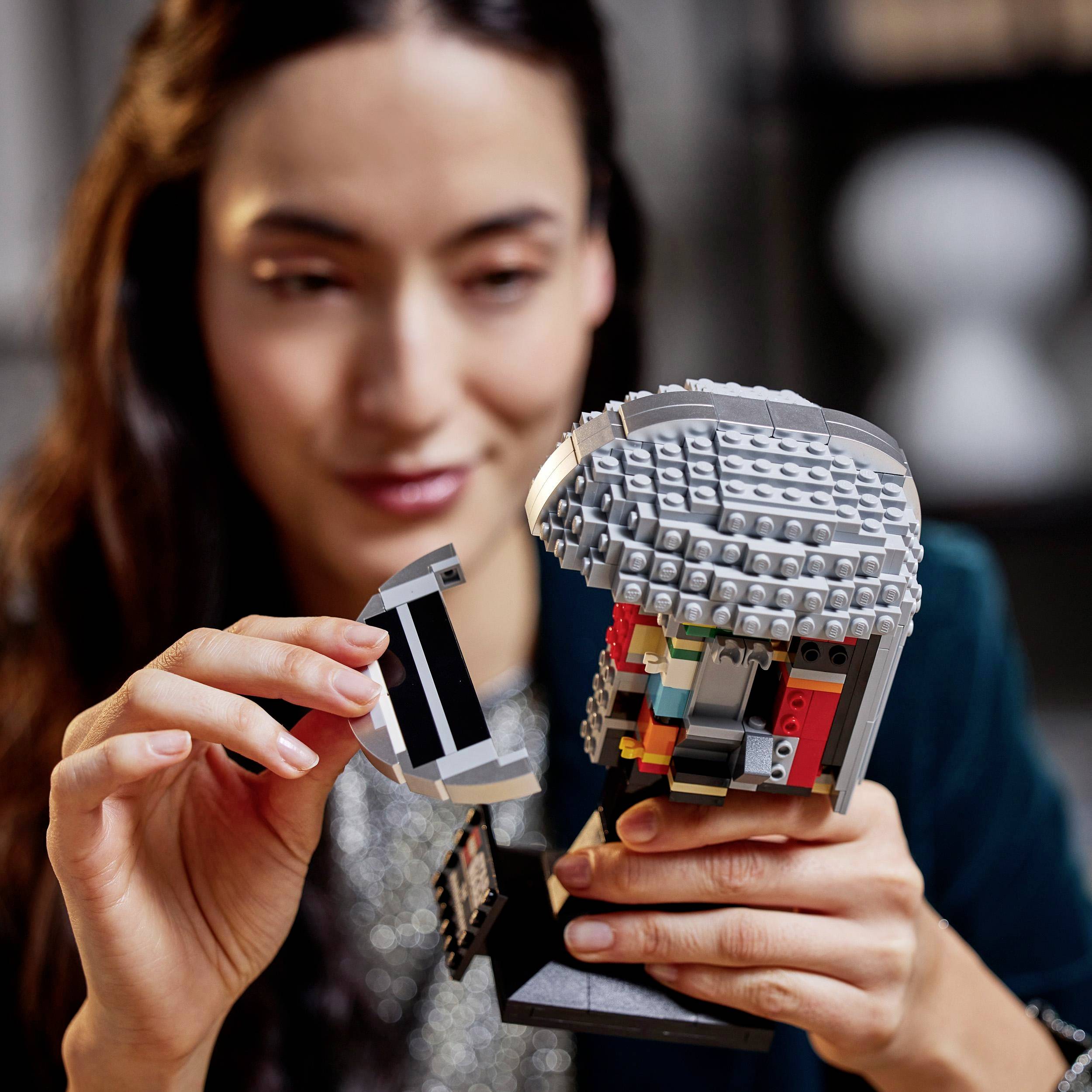 A person is holding a model constructed from building blocks that resembles a helmet. The focus is on the detail of the construction in an interior setting.