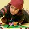A child is playing with colourful building blocks on a green surface. They are wearing a red cap and appear to be deeply concentrated.
