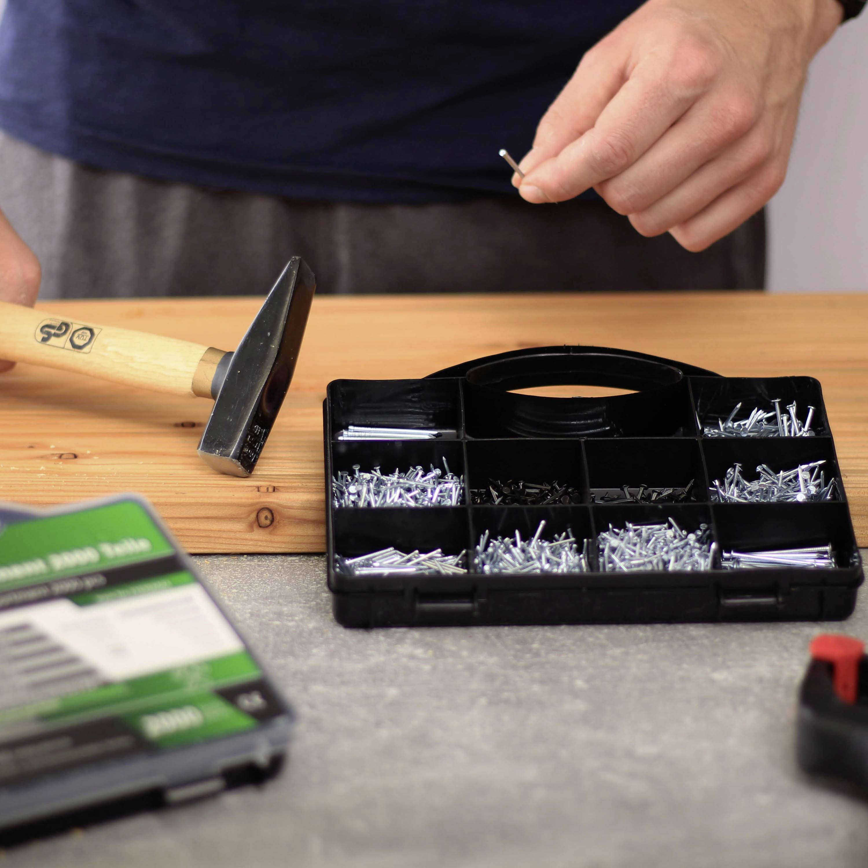 Someone is holding a nail, ready to hammer it. Next to them, an open box with various nails and screws lies on a table.