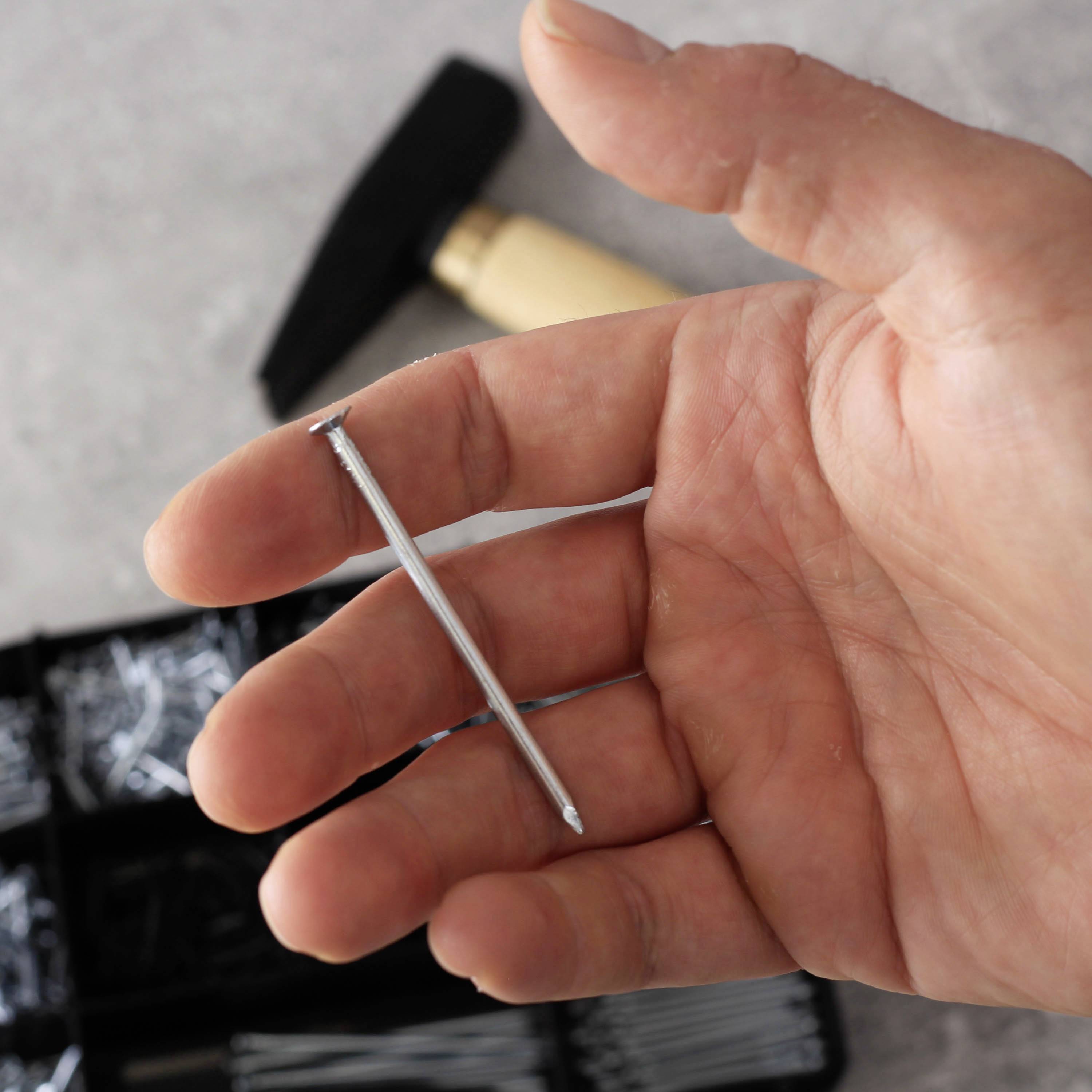 A hand holds a nail over a table with various nails and a hammer in the background.