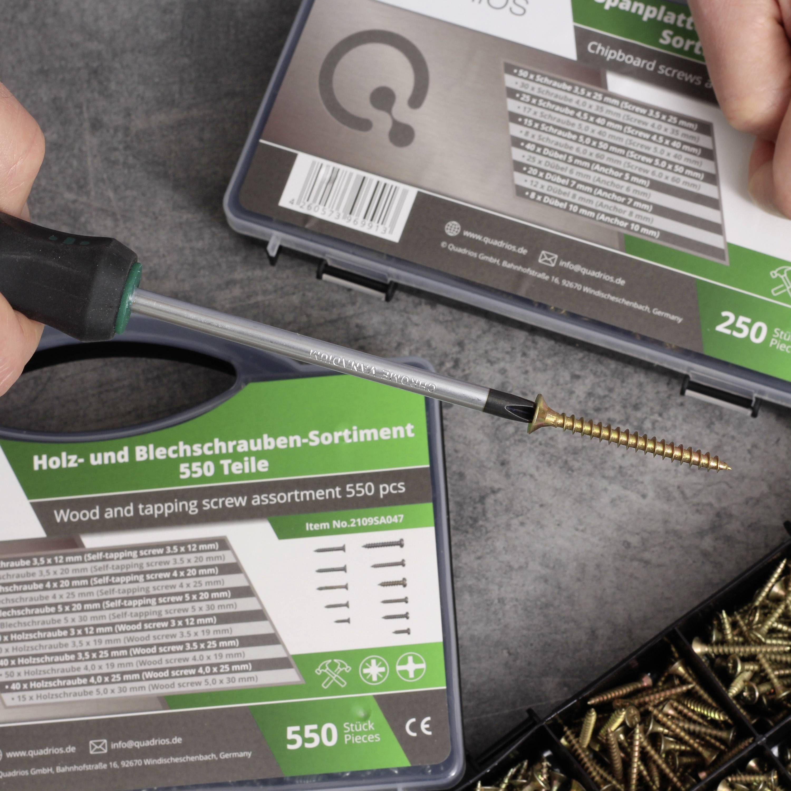 A hand holds a screwdriver with a long golden screw over a box containing wood and metal screws of various sizes.