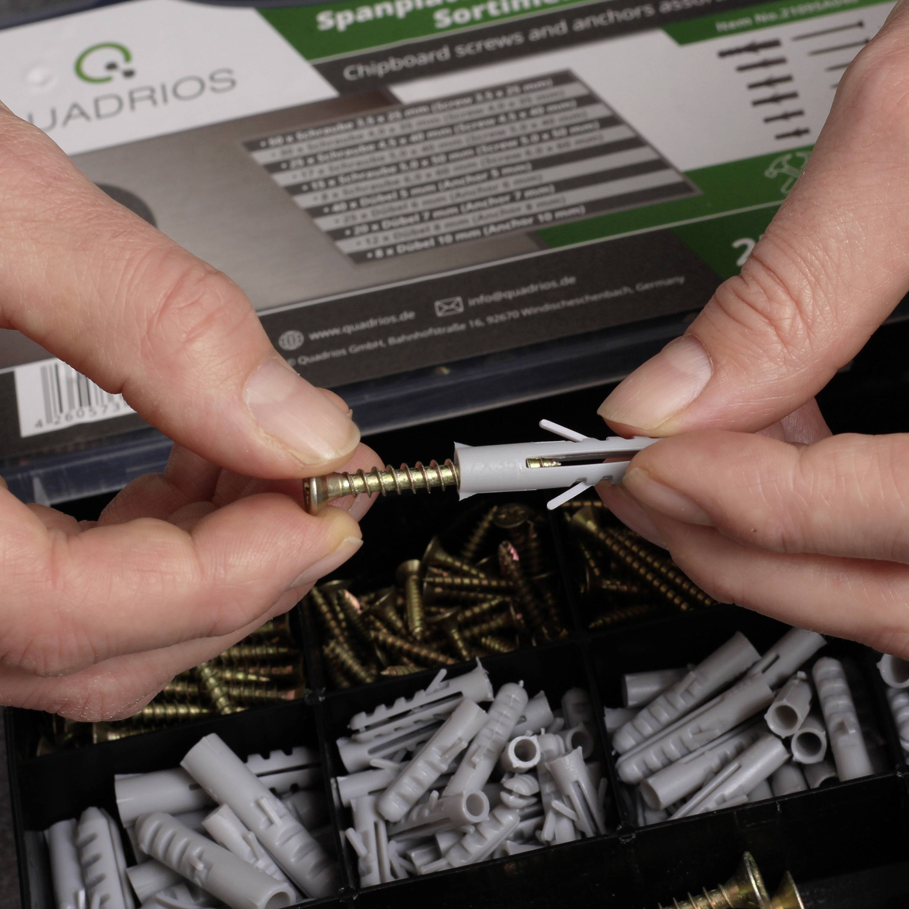 A hand is holding a screw and a wall plug, while a second hand points at them. In the background, a cardboard box with screws and wall plugs is lying.