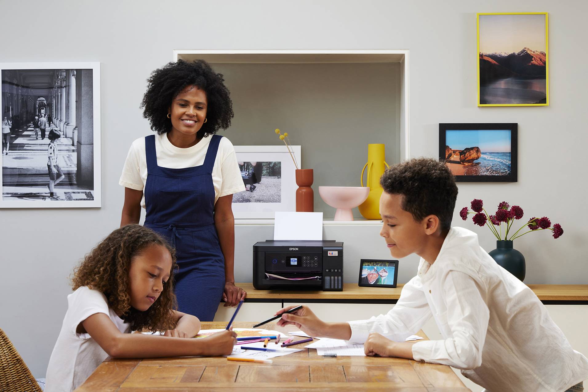 A smiling woman looks at two children painting together at the table. In the background, there are framed pictures and a printer.