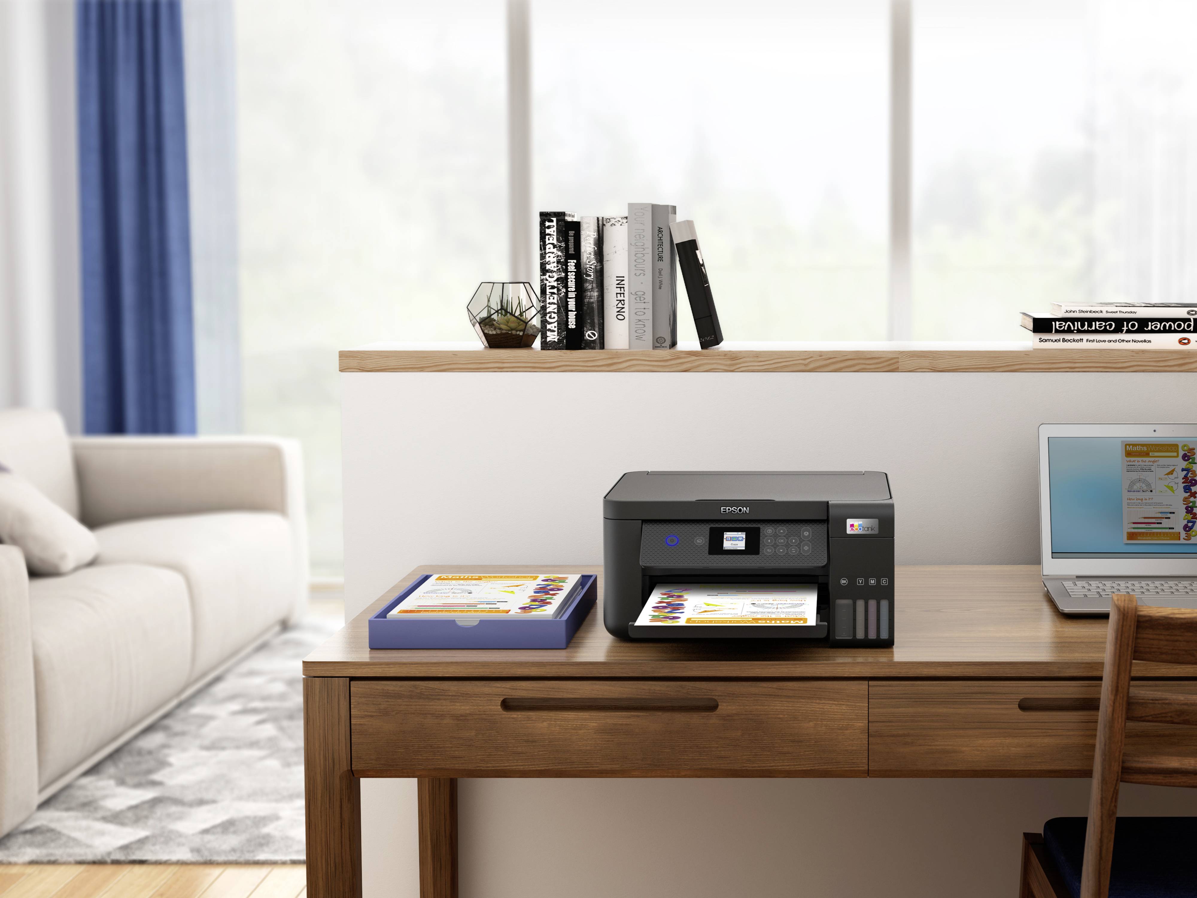 A printer on a wooden table is printing a coloured document. Books and a plant are in the background. A sofa is visible.
