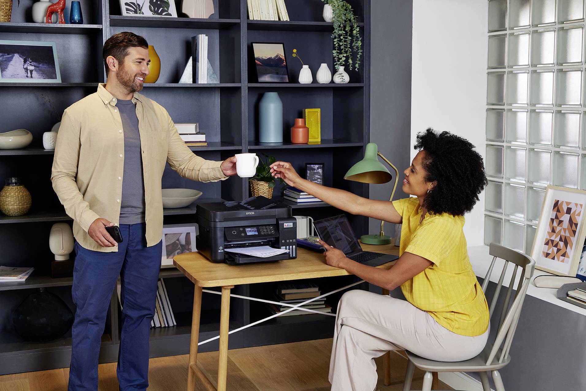 A man hands a woman a cup while she sits at a desk operating a printer. A bookshelf is visible in the background.