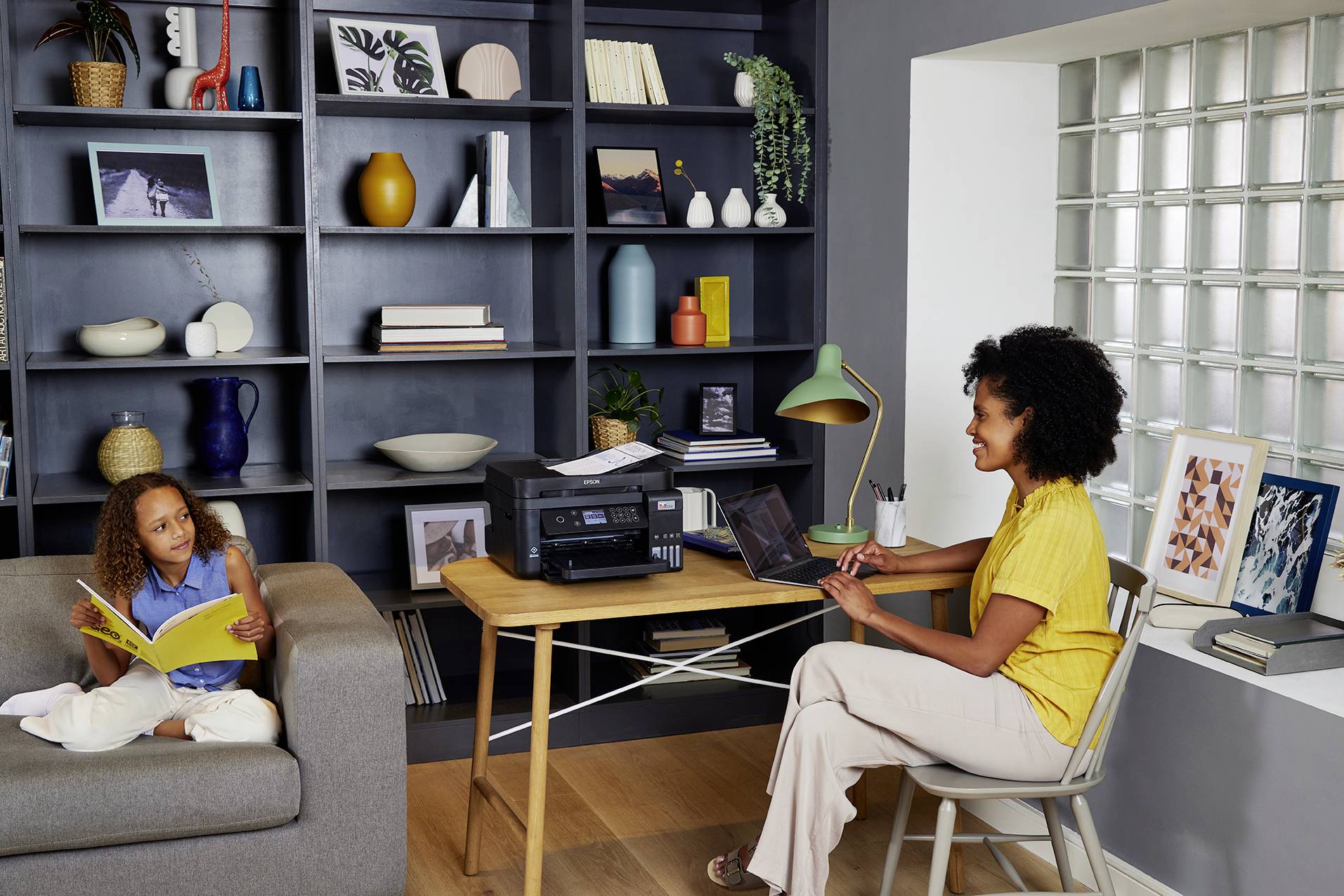 A woman is working on a laptop at a desk with a printer, while a child sits on the sofa reading a book. Bookshelf in the background.