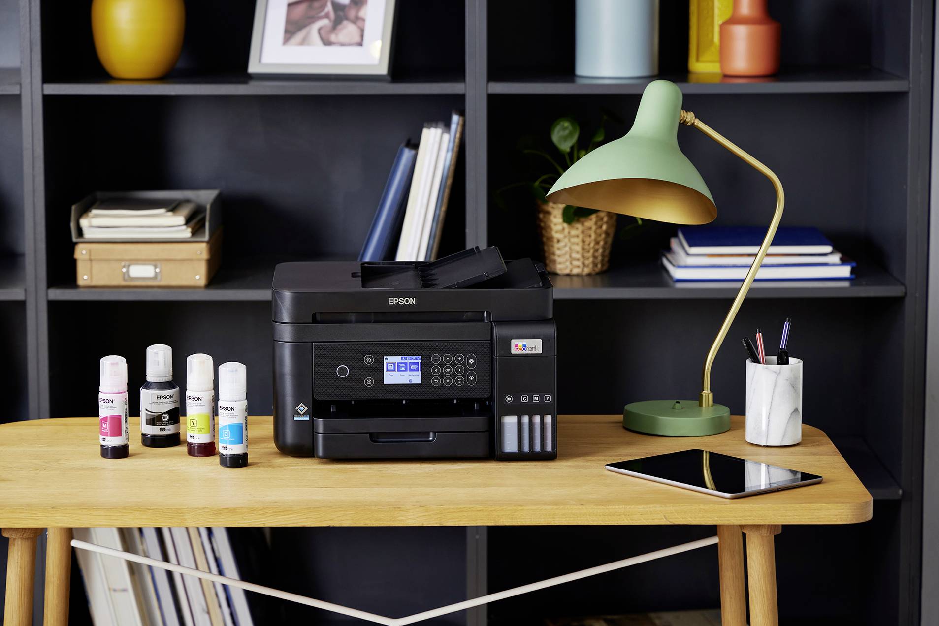 On a wooden table stands a printer with ink bottles beside it. In the background, a bookshelf and a green lamp are visible.