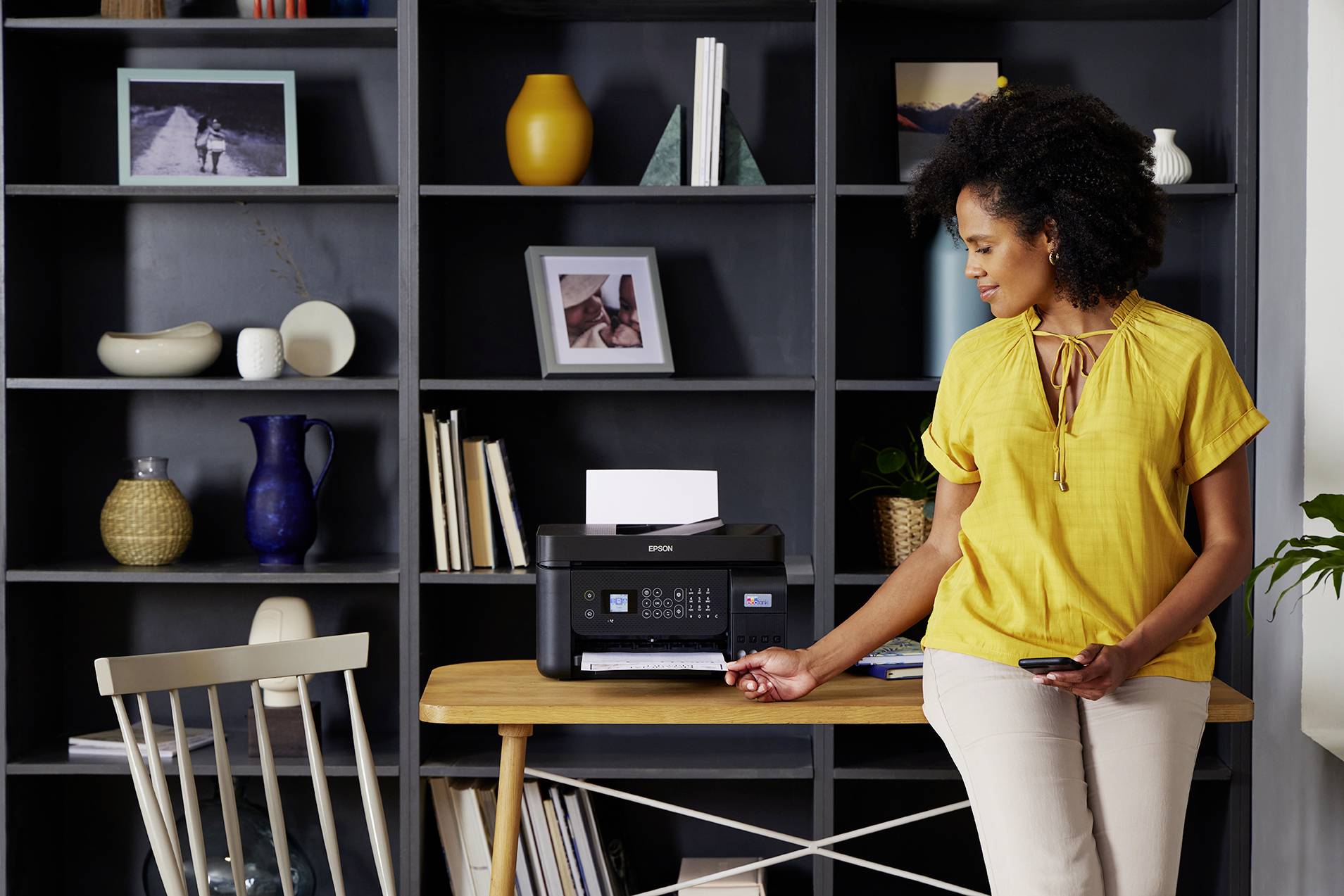 A woman in a yellow top is standing next to a printer on a table, holding a smartphone and looking at a printed page.