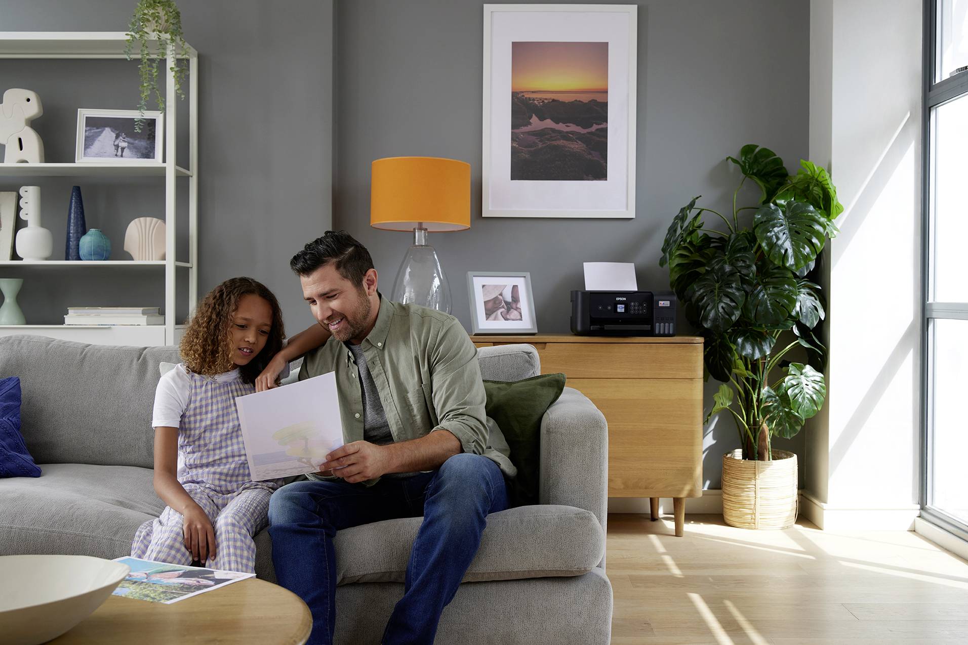 A smiling man sits on a sofa with a child, showing him a book. In the background, there is a bookshelf and a plant.